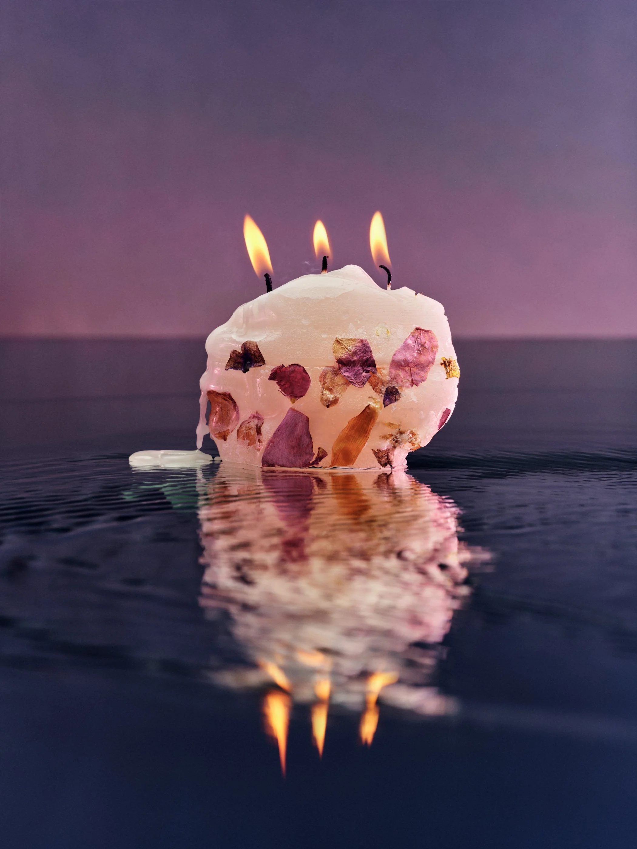 A birthday cake with three lit candles floating on water, with the cake's reflection visible on the water surface.