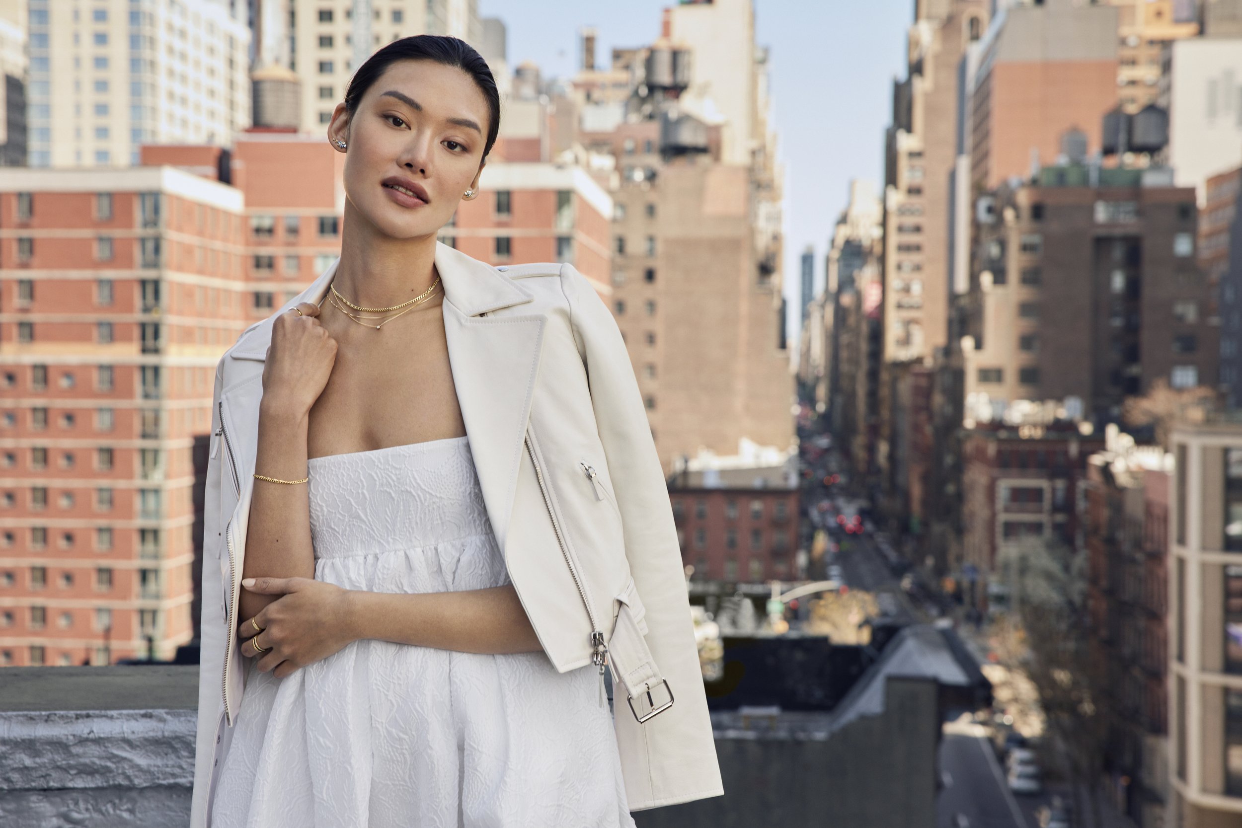 A woman standing on a rooftop with a cityscape background, dressed in a white strapless dress, white jacket, and jewelry.