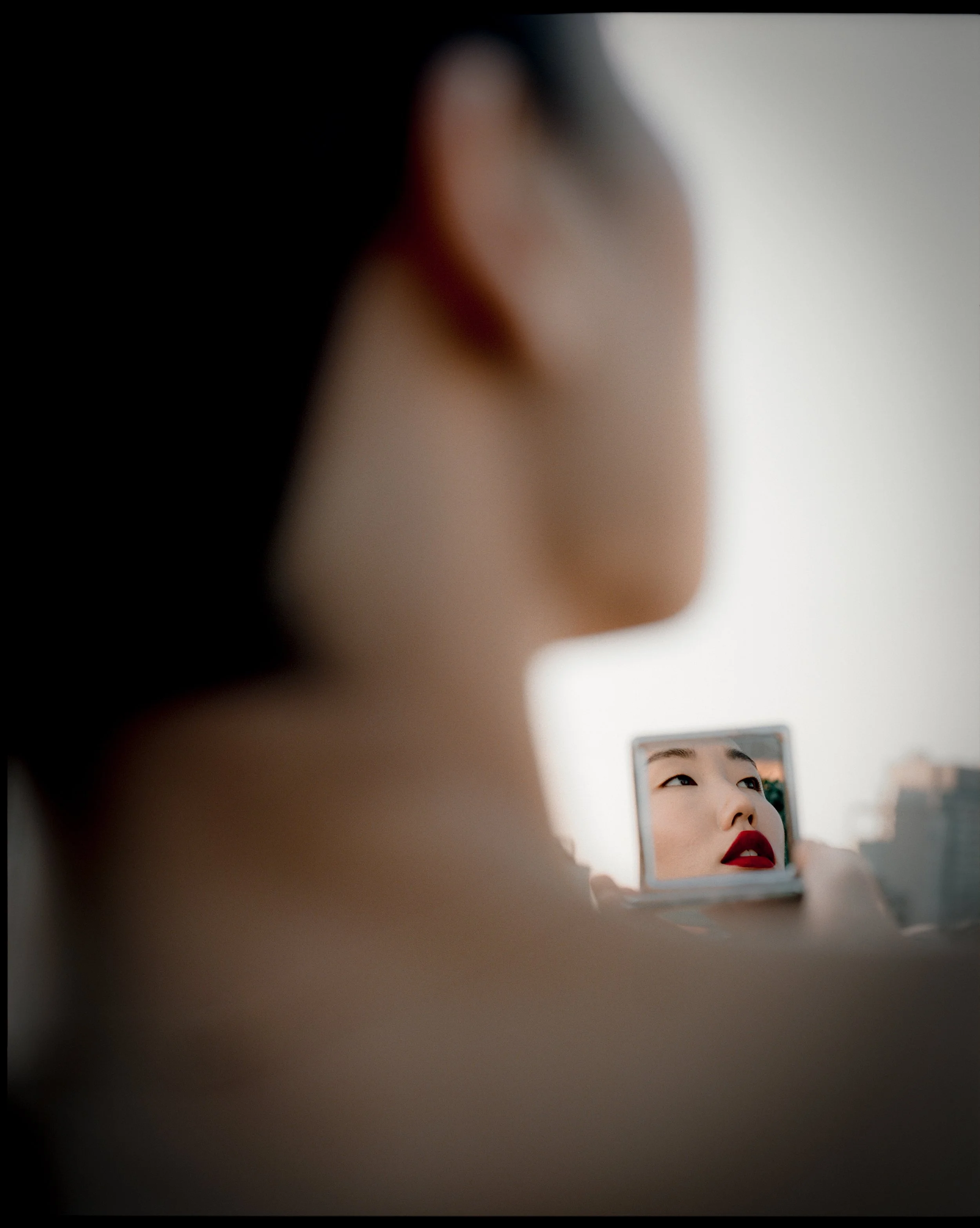 A woman applying red lipstick while looking into a small mirror, with her reflection showing her face with red lipstick and makeup.