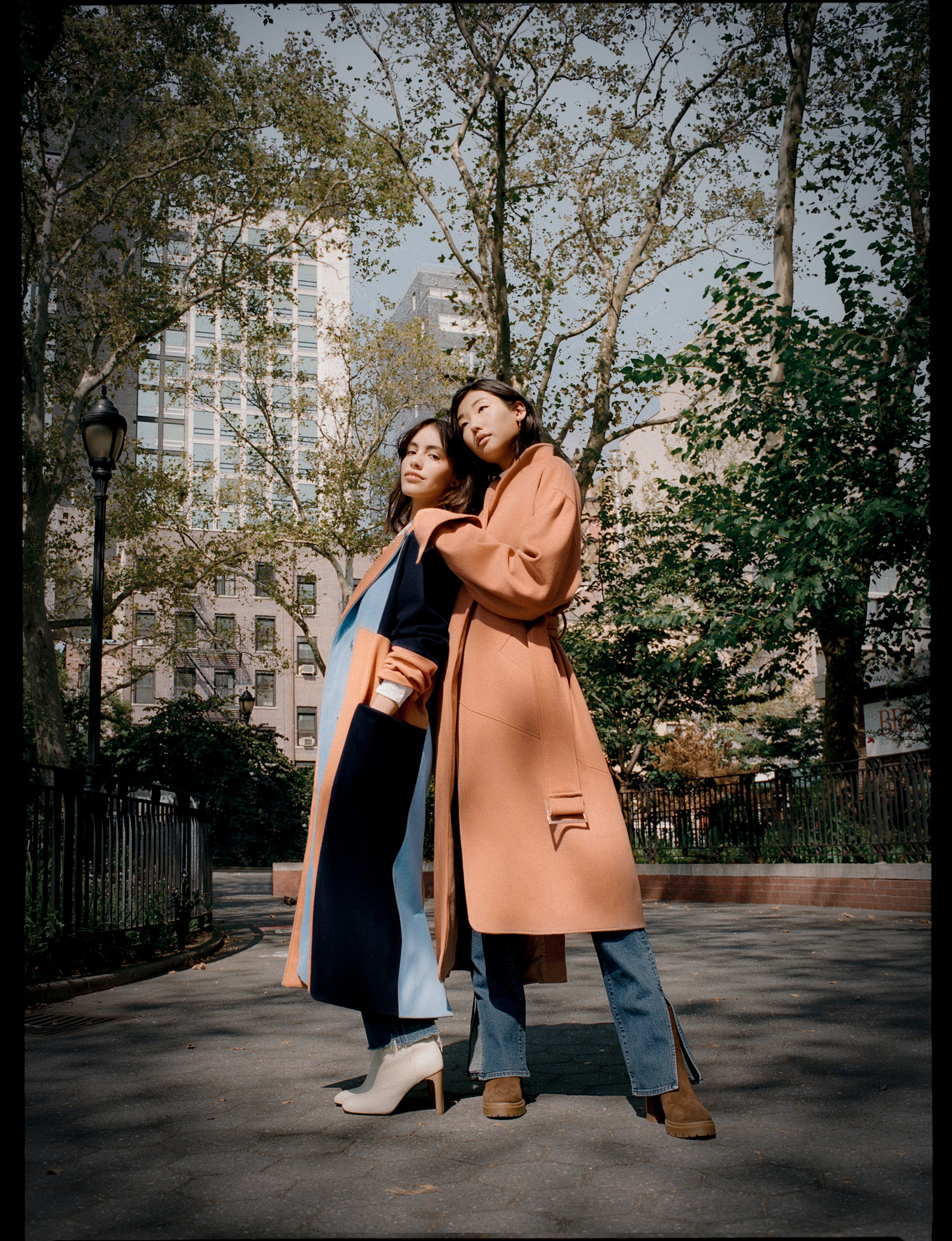 Two women in stylish coats and boots standing close together in a city park with trees and tall buildings in the background.