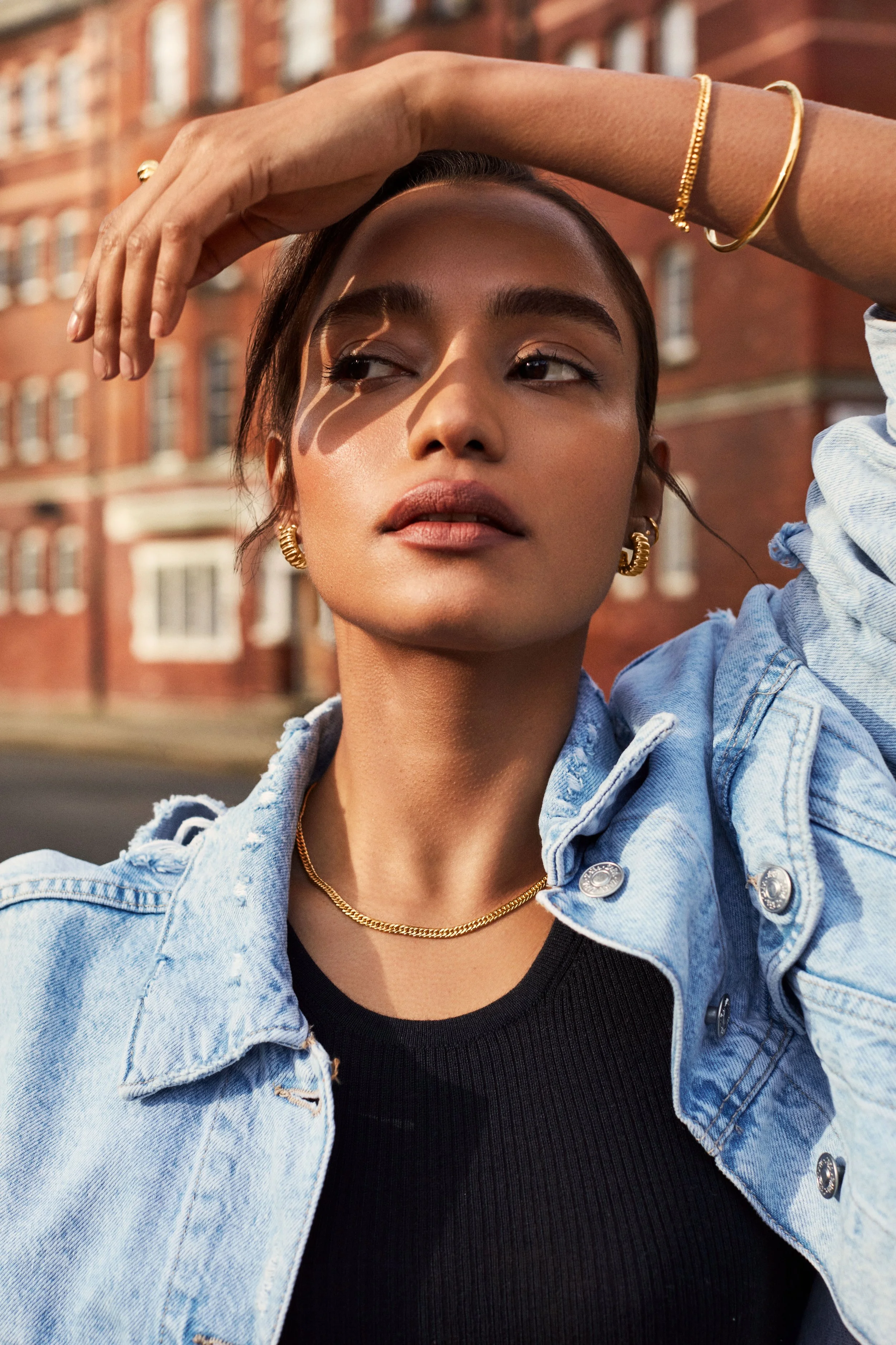 A woman wearing a denim jacket, gold jewelry, and a black top, standing outdoors with a brick building in the background. She has her arm raised over her head, casting a shadow on her face.