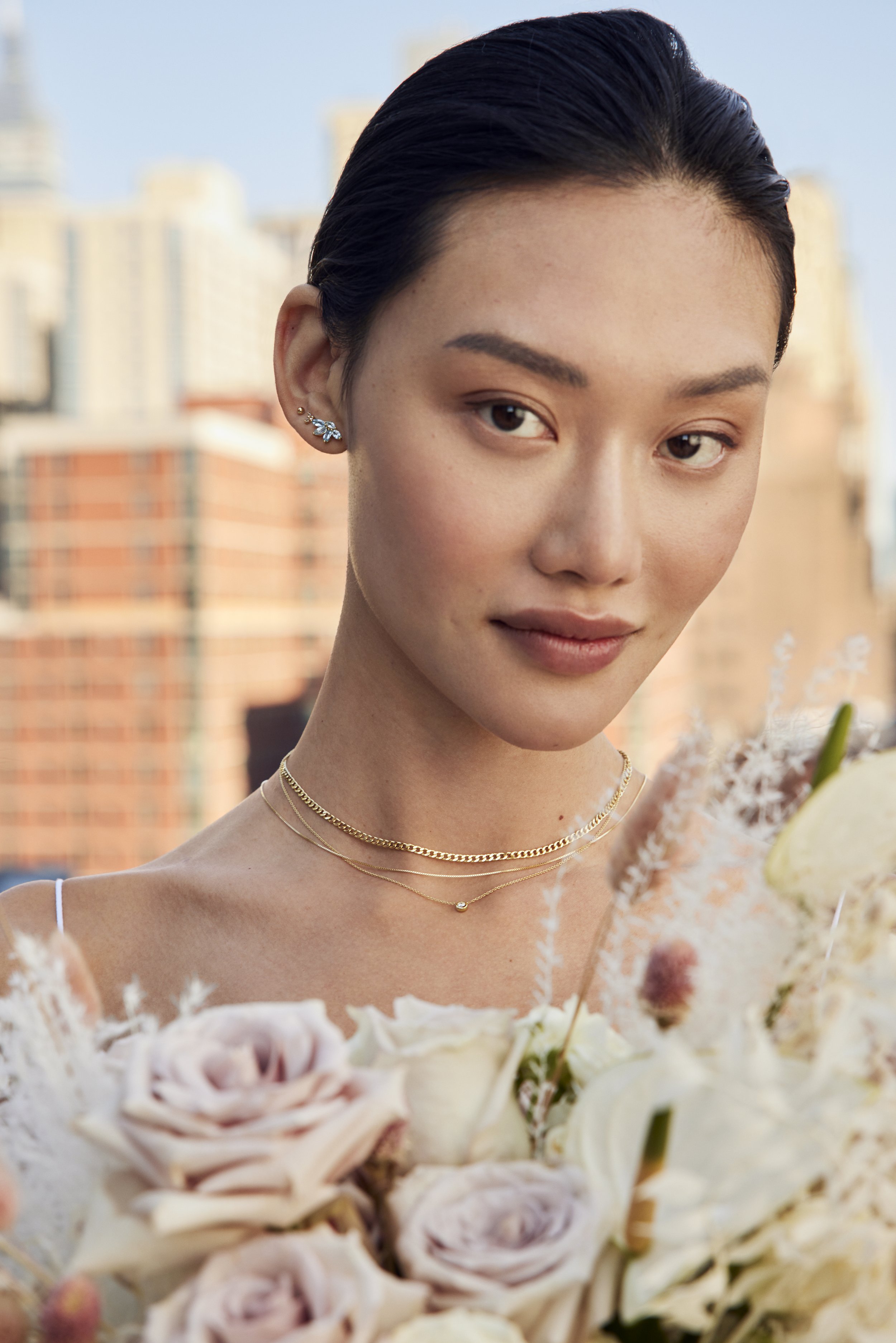 A young woman with dark hair, wearing jewelry, holding a bouquet of flowers, with city buildings in the background.