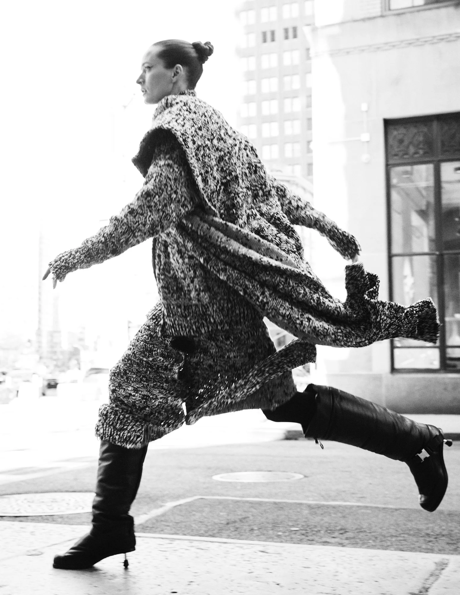 A woman in a patterned coat and tall boots running outdoors in an urban setting.