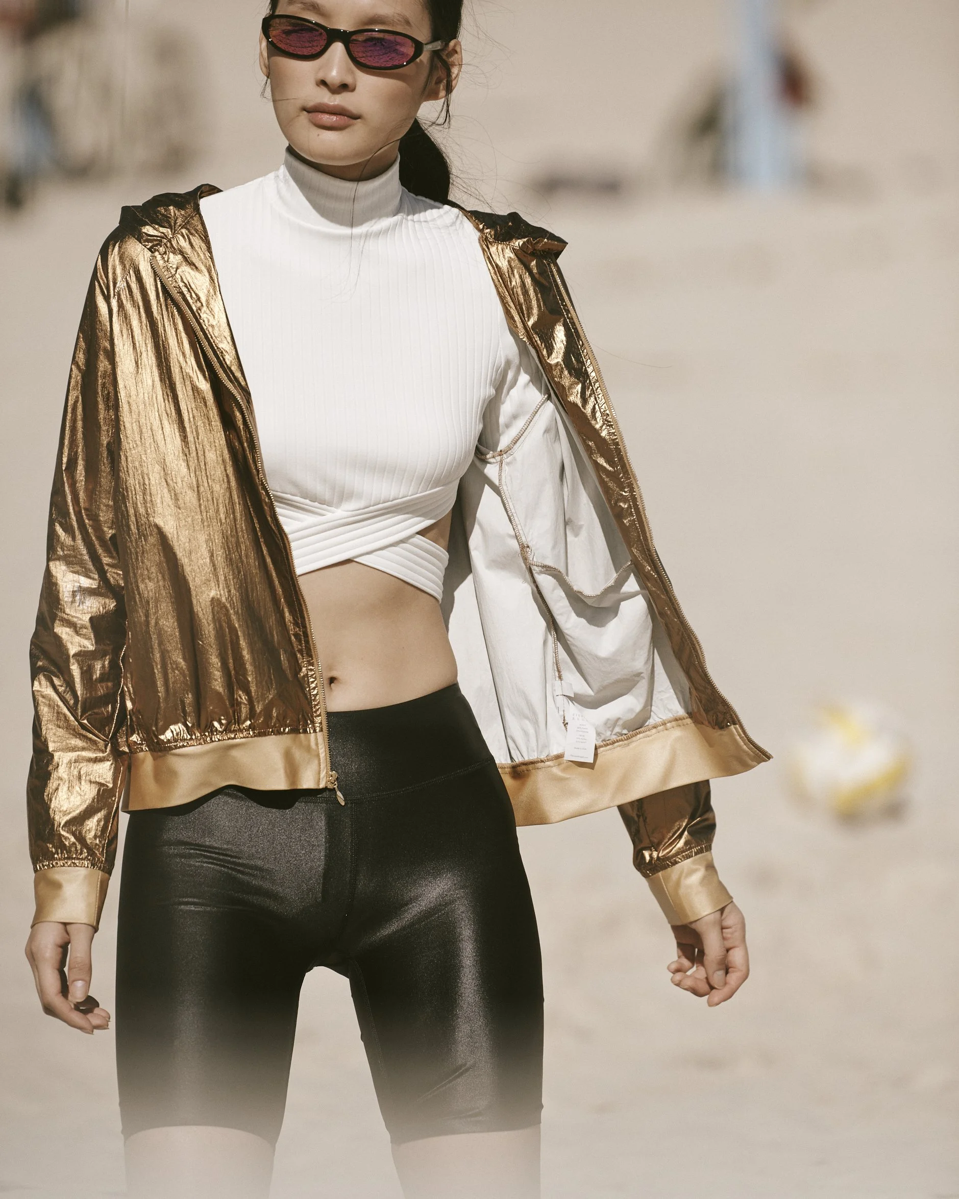 Woman wearing sunglasses, white cropped top, gold jacket, and black shorts on the beach.