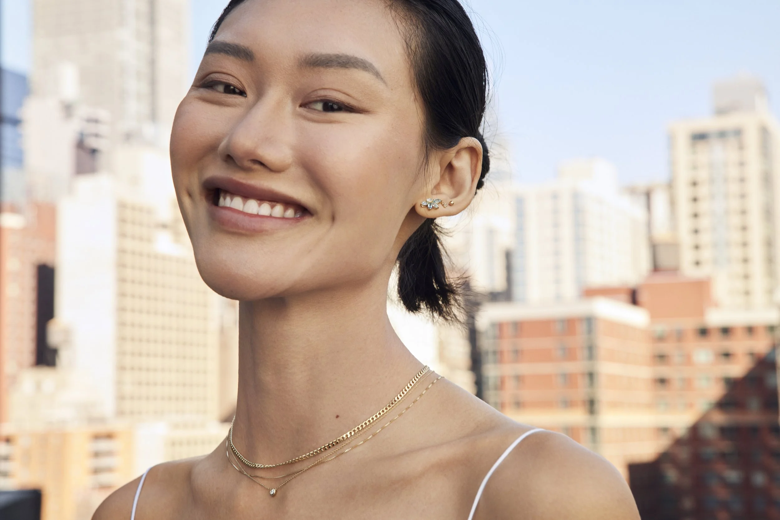 A young woman smiling outdoors in a city setting with tall buildings in the background.