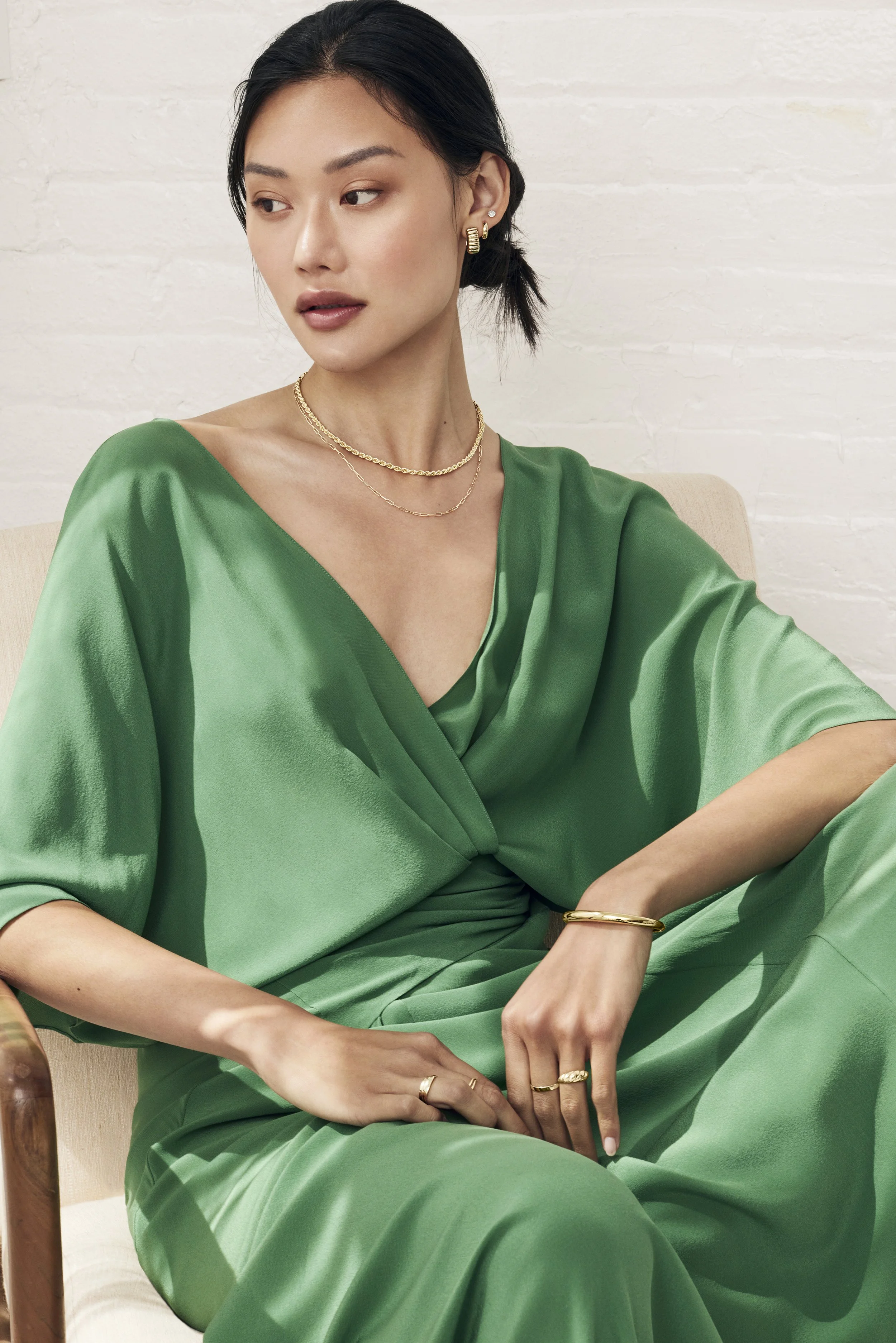 A woman wearing a green satin dress sitting on a beige chair, accessorized with gold jewelry including earrings, necklaces, rings, and bracelets, with a white brick wall in the background.