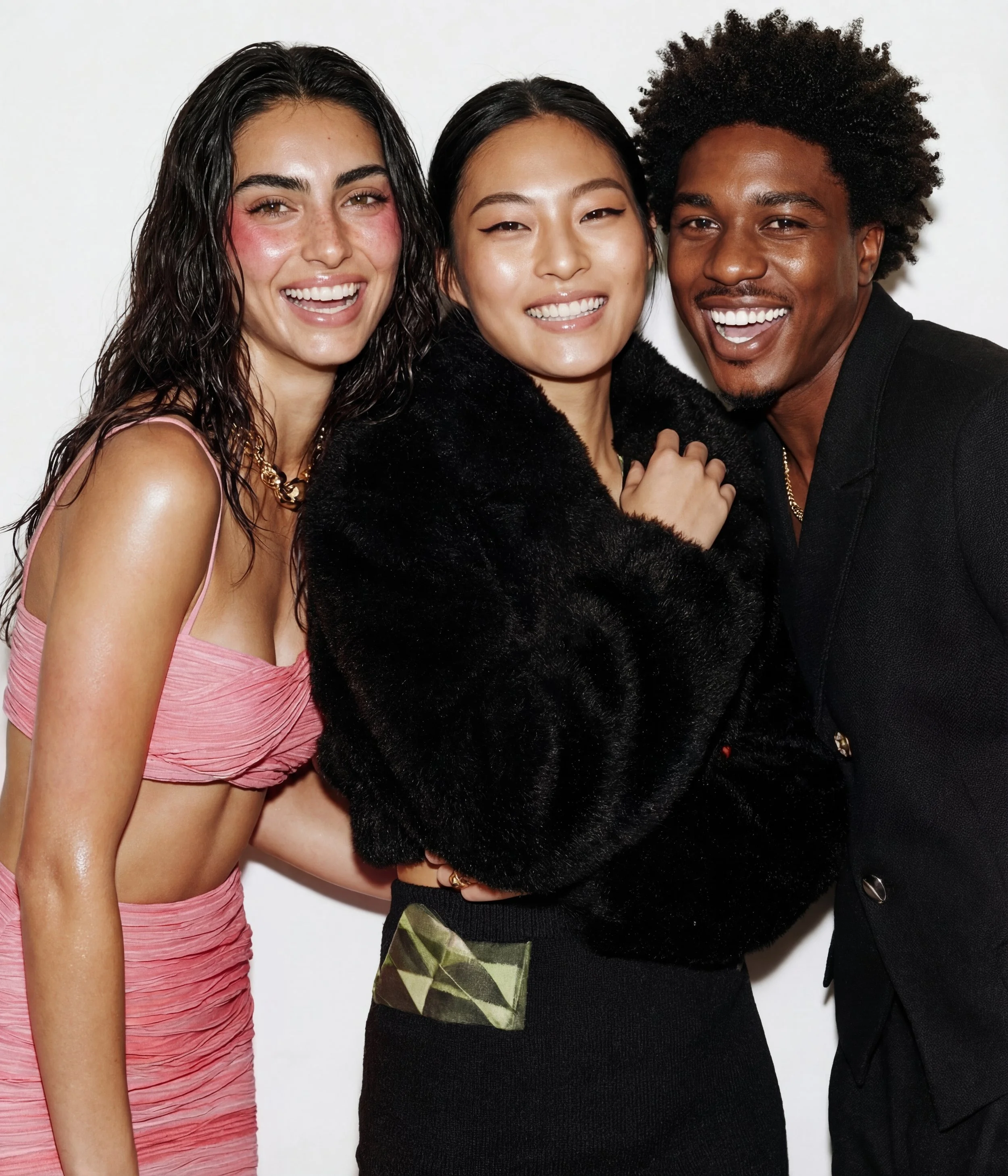 Three smiling young adults, two women and one man, posing together. The woman on the left has long, dark, wavy hair and is wearing a pink ruched crop top. The woman in the center has straight, dark hair and is wrapped in a black faux fur coat. The ma