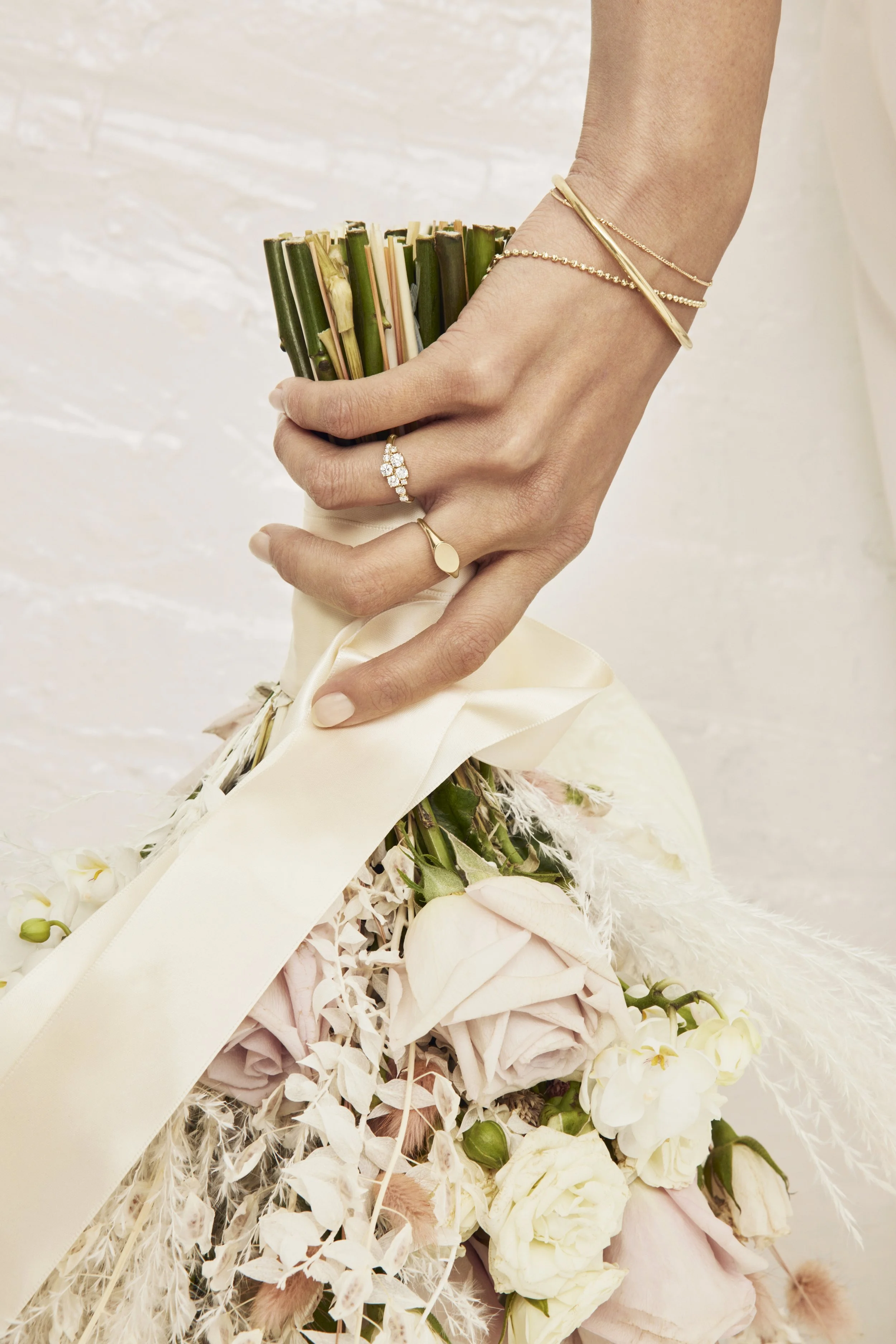 Close-up of a woman's hand holding a bouquet of pale pink, cream, and white flowers, with gold jewelry including rings and bracelets.