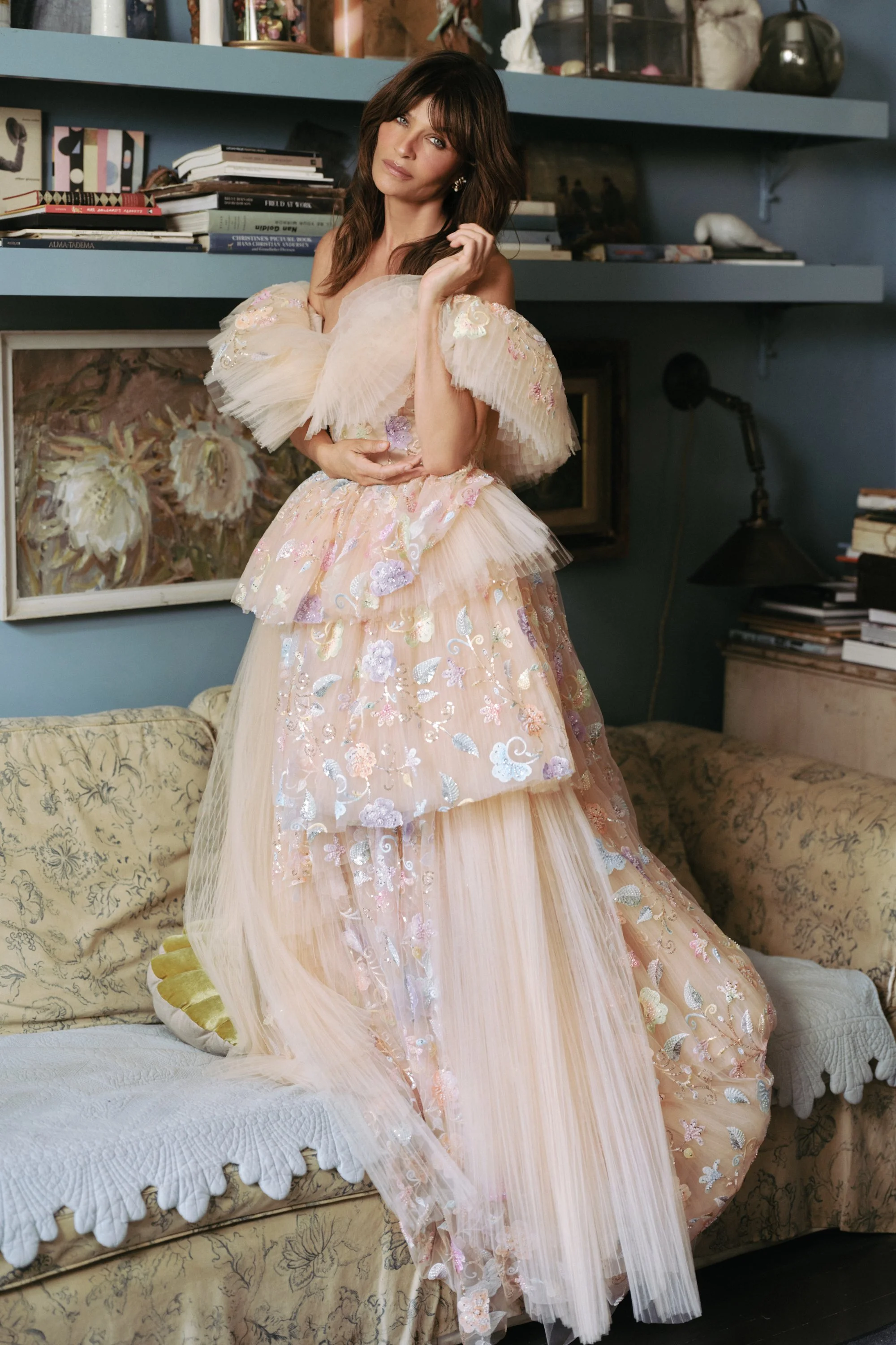 A woman in an elaborate, pastel-colored floral dress with puffed sleeves, standing on a vintage sofa in a cozy, art-filled room.