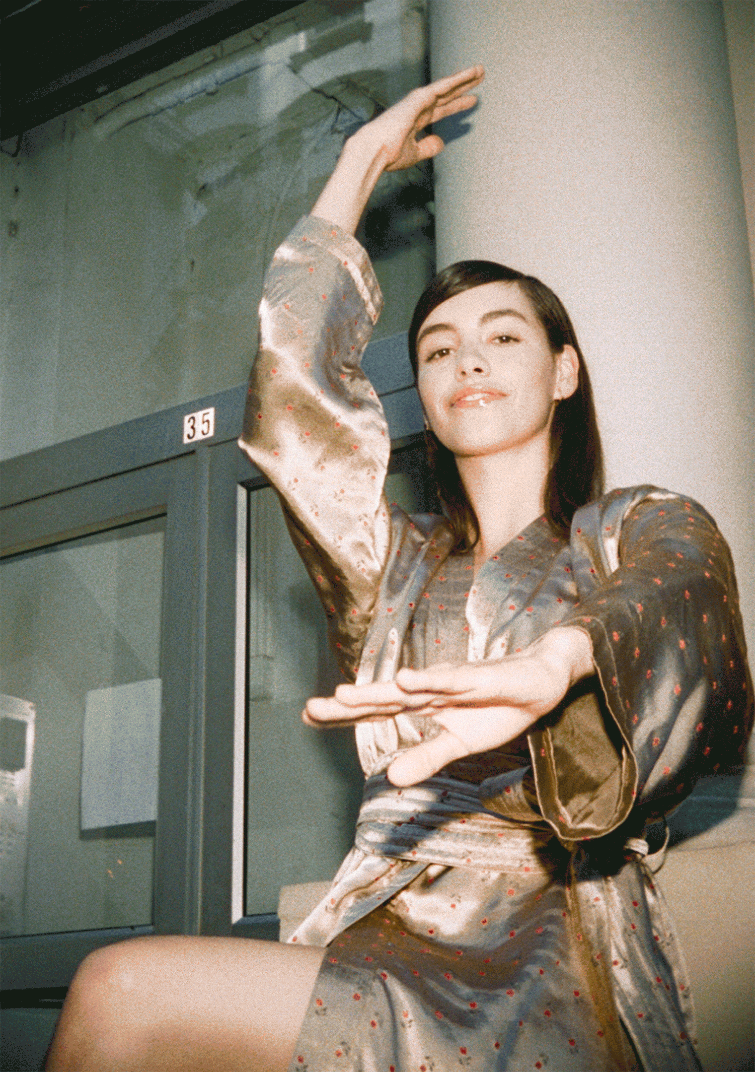 A young woman with dark hair and a light complexion, dressed in a shiny, patterned silk robe, sitting on a bench beside a glass-fronted public telephone booth. She is looking at the camera with a subtle smile, her right arm raised with her hand near 