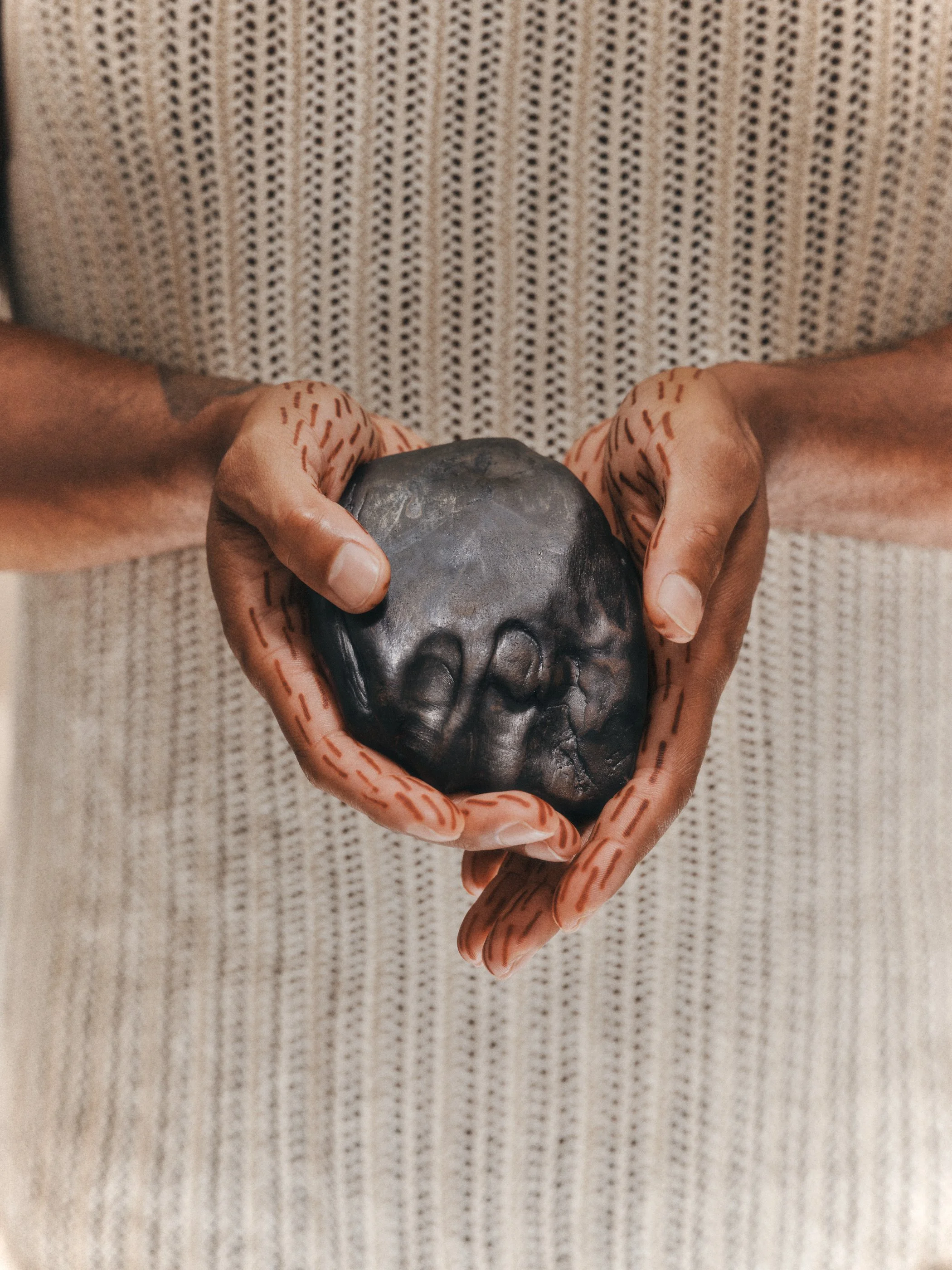 Person holding a black stone in their hands, with markings on their fingers, in front of a beige, textured background.