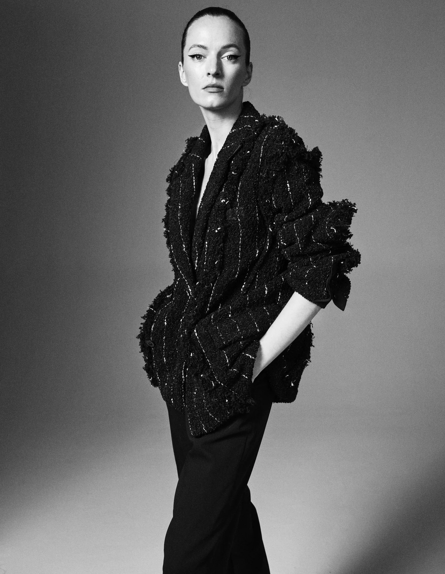 A woman with her hair pulled back, wearing a textured, striped jacket and dark pants, standing with one hand in her pocket against a plain background.