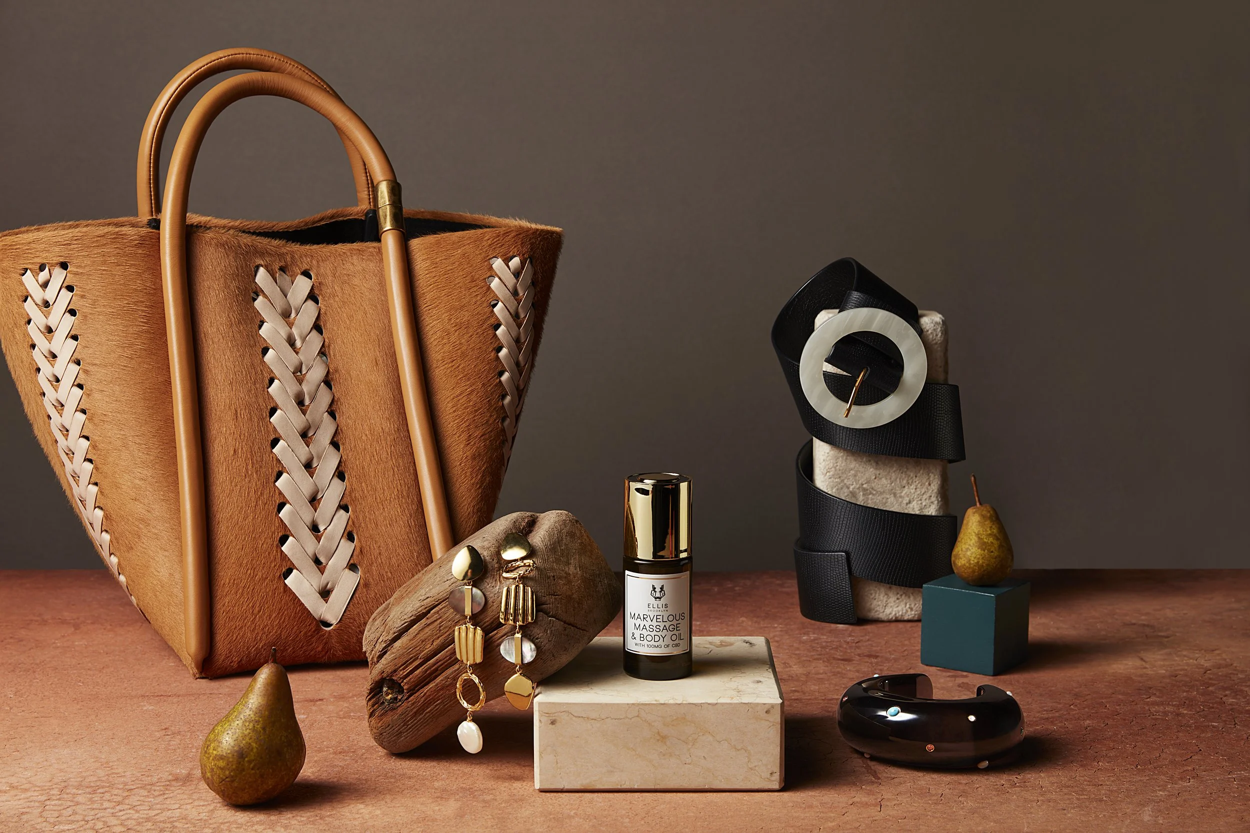 Stylish accessories on a wooden surface, including a large woven leather handbag, a wooden jewelry holder with dangling earrings, a small bottle of massage oil, a black cuff bracelet, decorative pears, and geometric blocks, with a neutral background.