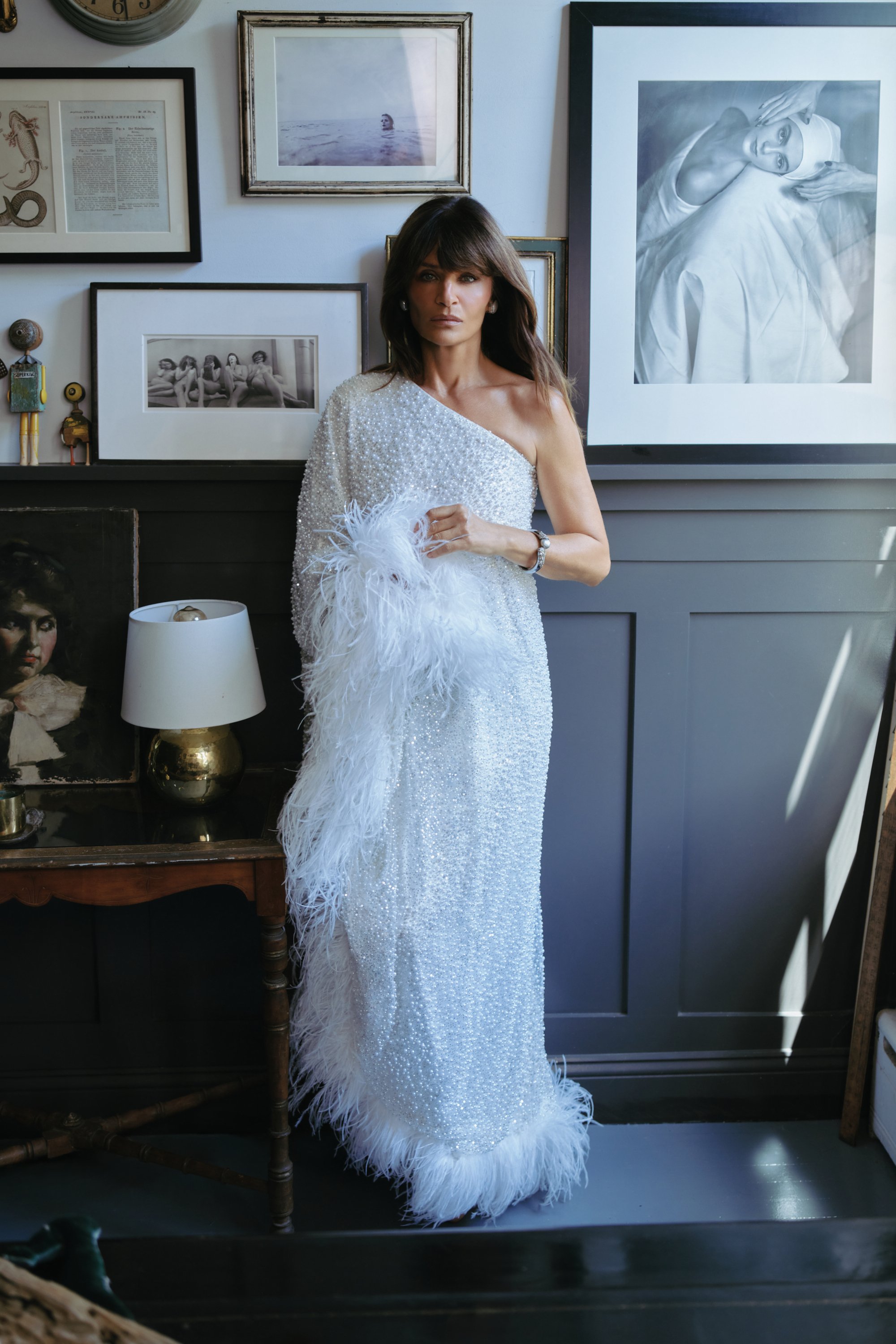 Woman in a one-shoulder, white, beaded dress with feather details, standing in a gallery room with framed art, photographs, and a lamp.