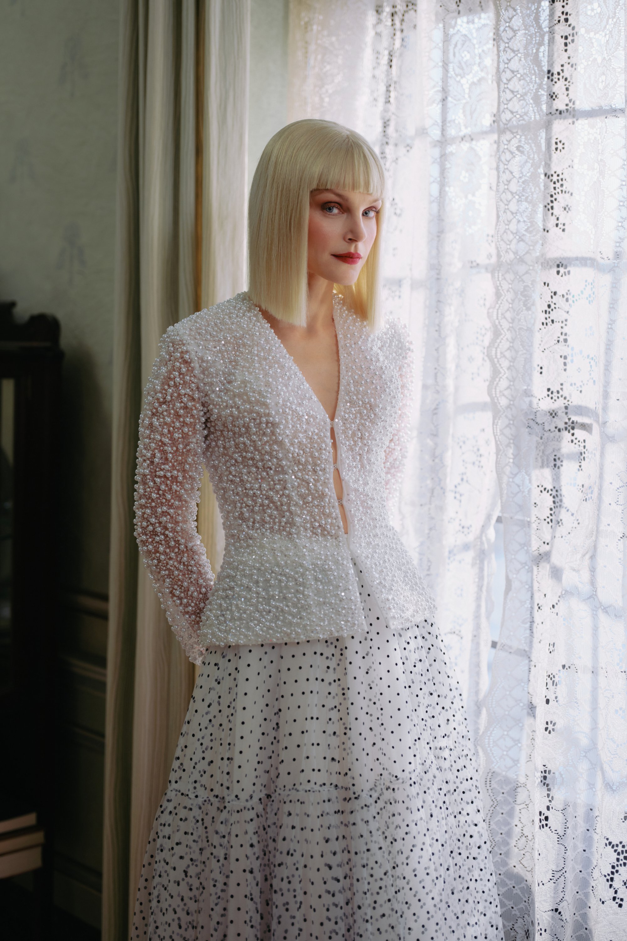 A woman with blonde hair in a straight bob hairstyle wearing a white, pearl-embellished jacket and a black polka dot dress, standing indoors near a lace curtain.