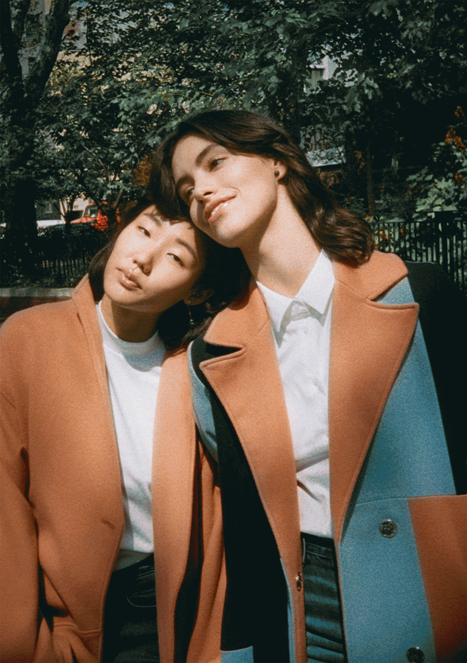 Two women standing close together outdoors, with trees and a fence in the background. One woman has short dark hair and the other has shoulder-length brown hair, both wearing white shirts and tan coats.