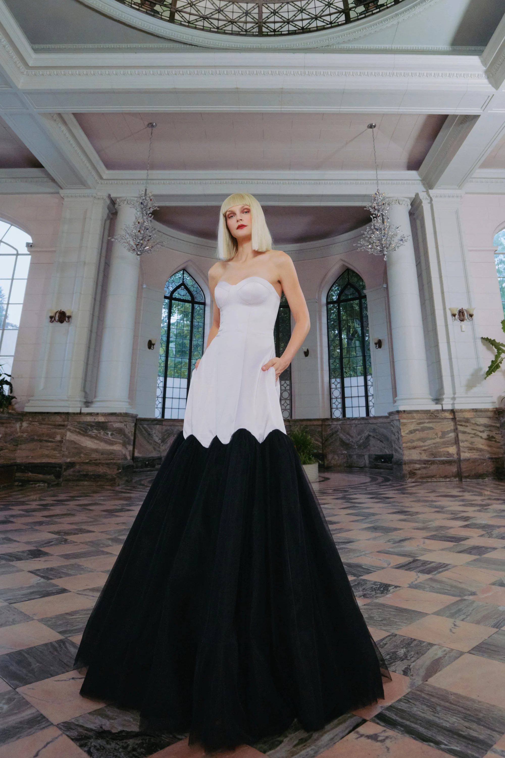 A woman with blonde hair wearing a strapless black and white gown in an ornate hall with high ceilings, chandeliers, and large arched windows.