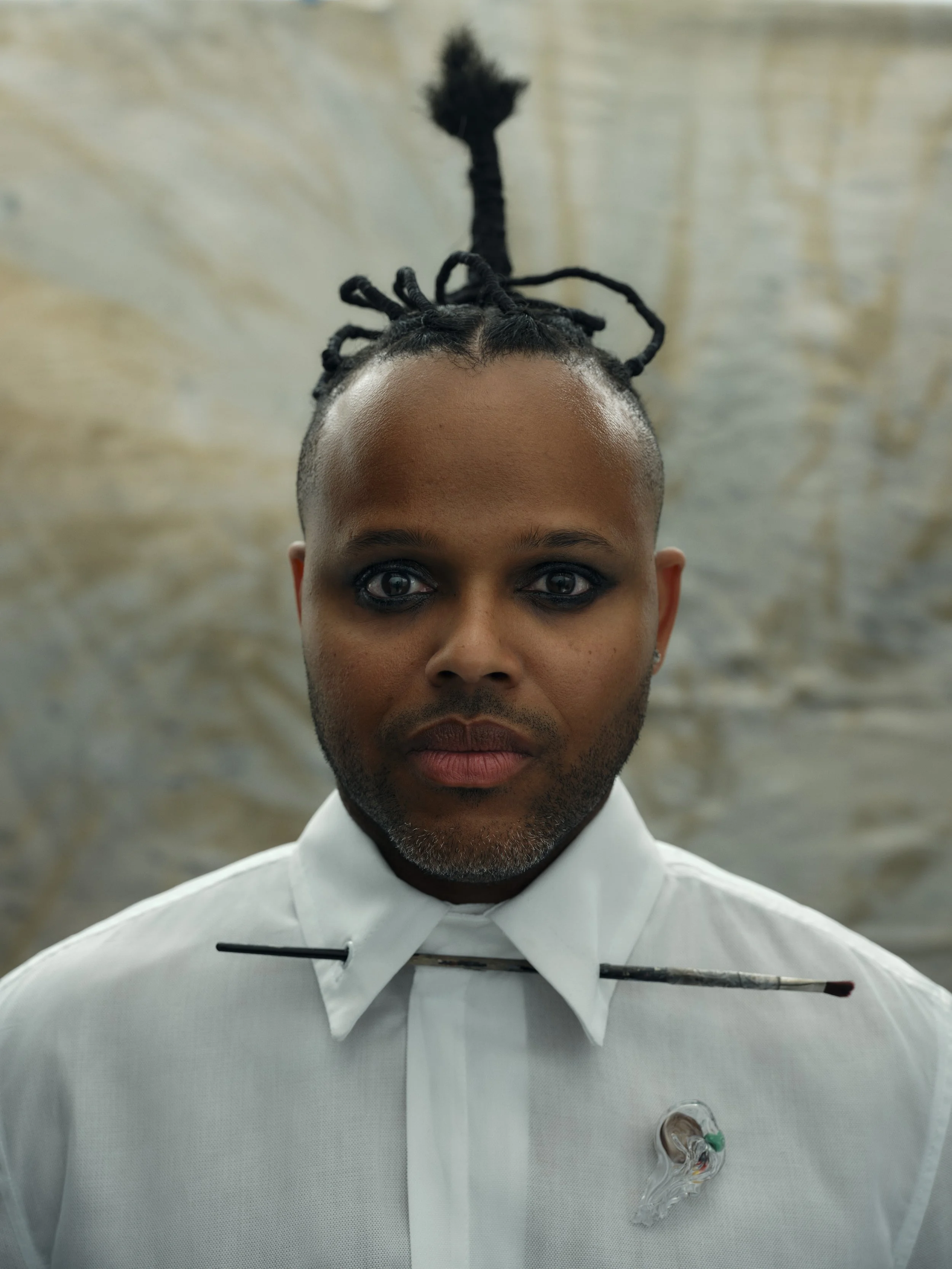 A person with a shaved head and dreadlocks styled on top, wearing a white collared shirt, a hairpin, and a gas mask hanging from the shirt with one strap.