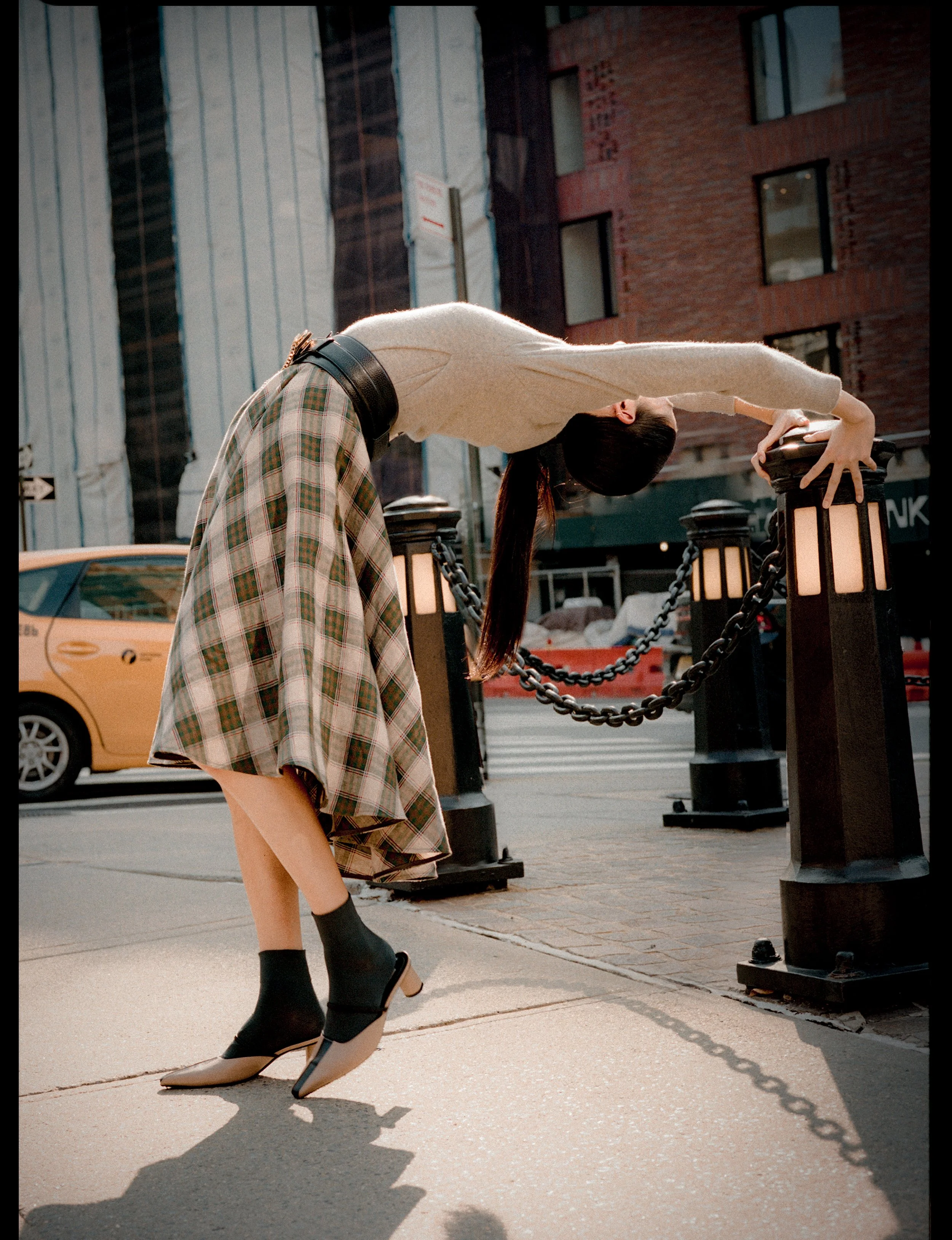 A woman in a beige sweater, plaid skirt, and high heels leans back over a railing on a city street.