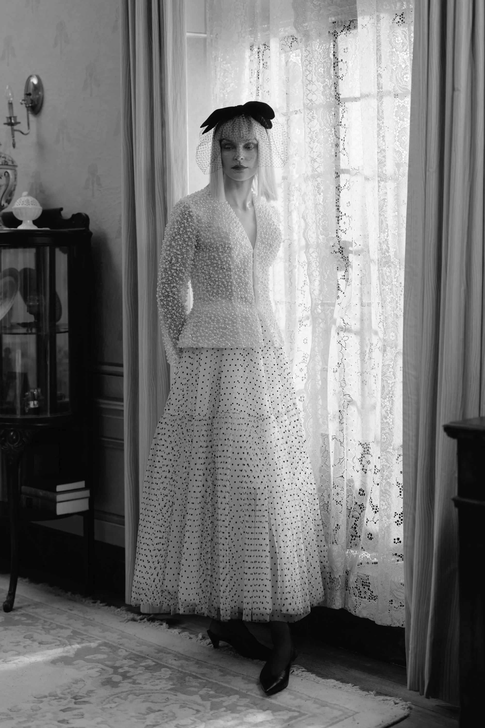A woman standing indoors by a window with lace curtains, dressed in a vintage-style, polka-dot dress, wearing a hat with a veil, and black high heels. The room has antique furniture and decorative items.