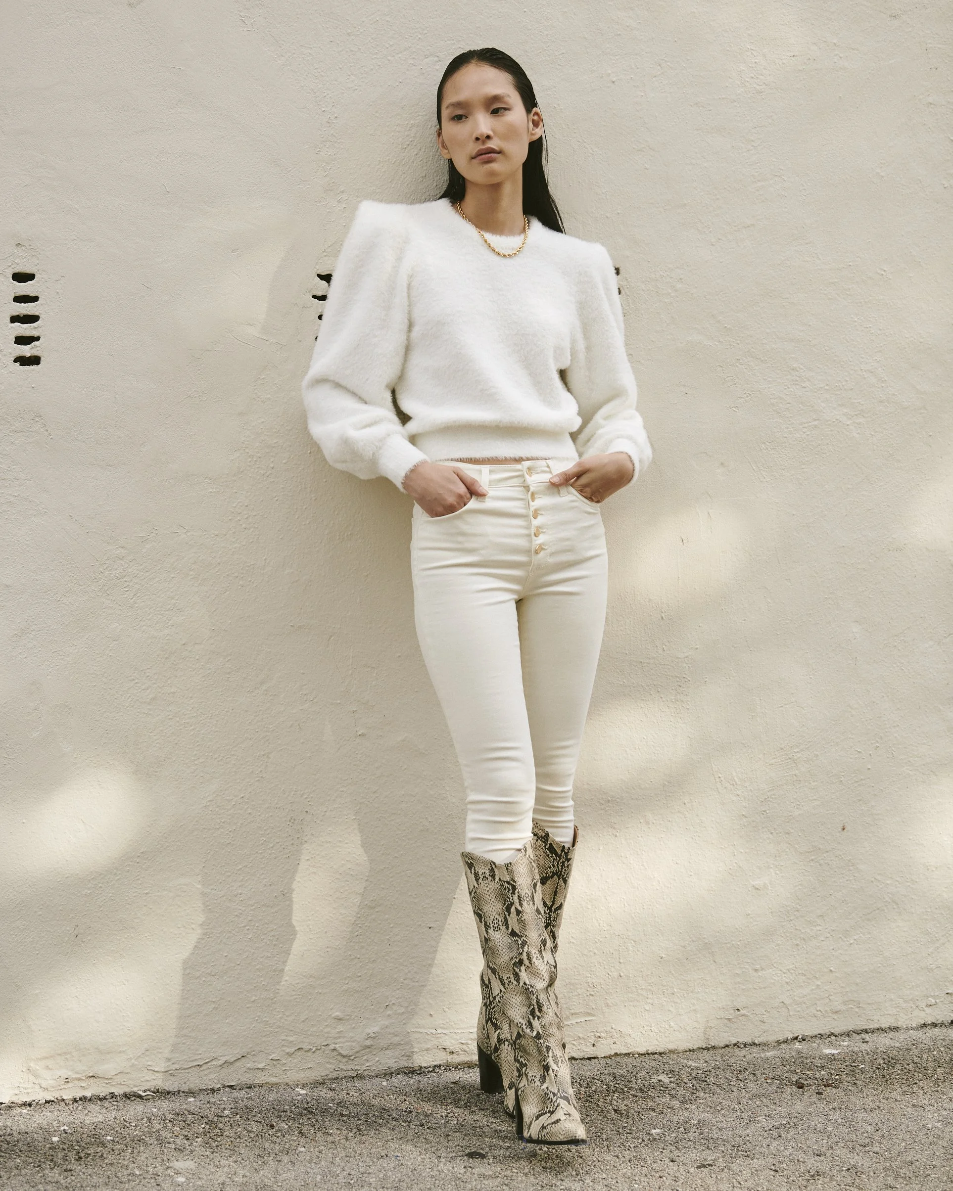 Woman wearing a white fuzzy sweater, white high-waist pants, and snake-print knee-high boots, standing against a beige wall.