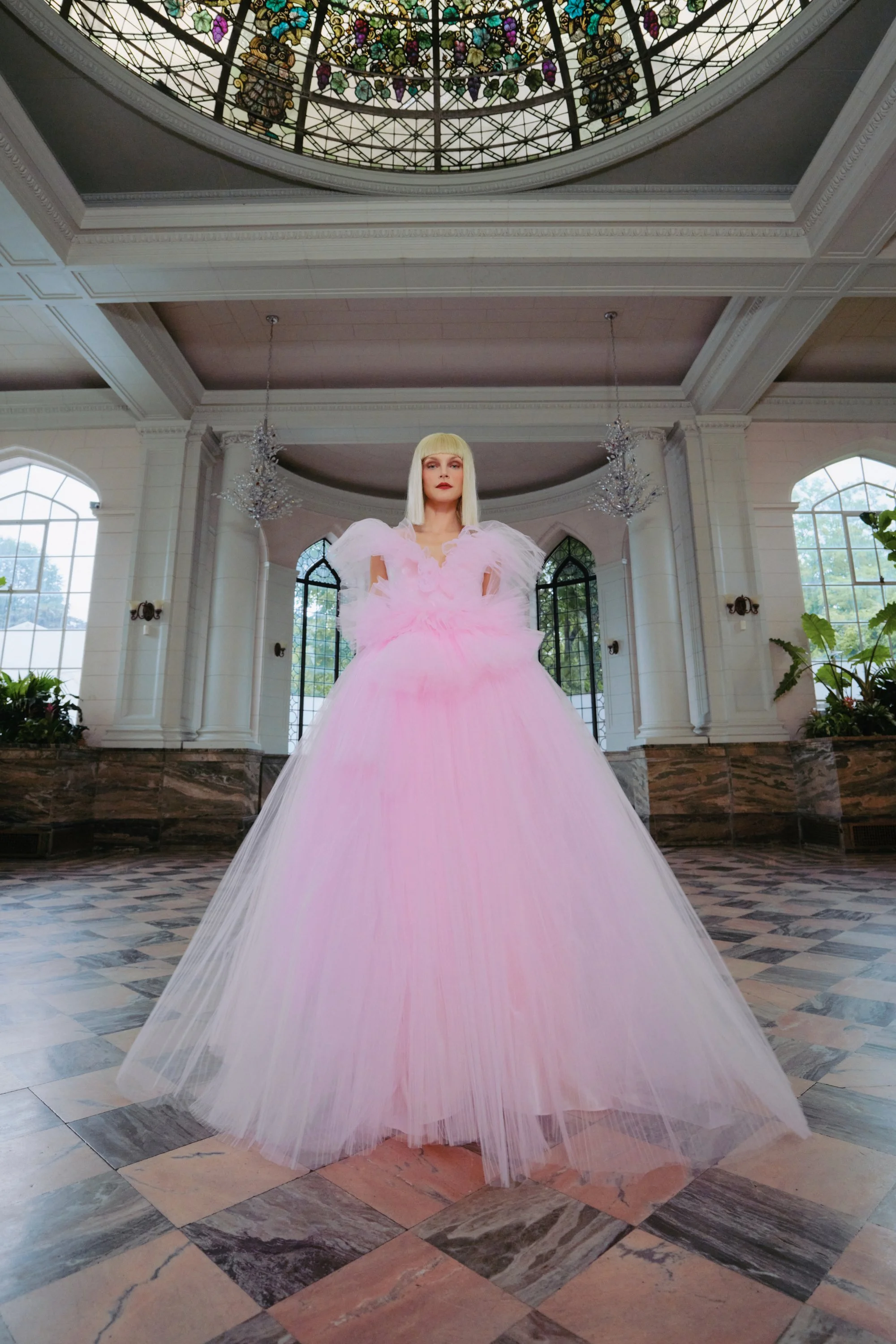 A person wearing a flowing pink gown with tulle details in a grand hall with tall windows, chandeliers, and a stained glass ceiling.