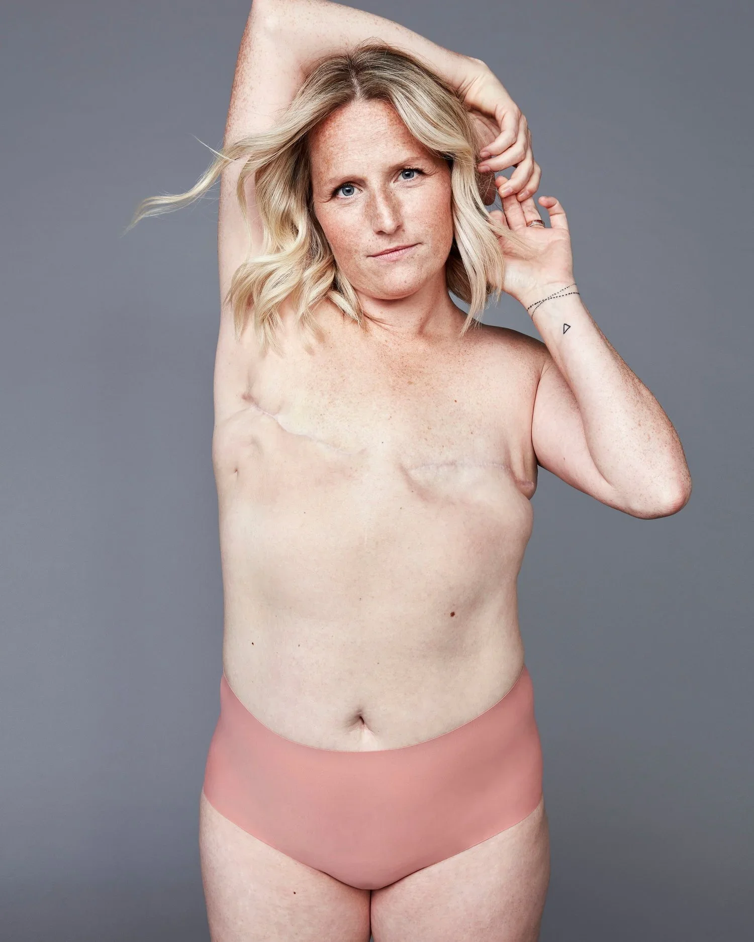 Portrait of a woman with blonde hair, freckles, and blue eyes, standing against a gray background. She is topless, wearing pink underwear, with one arm raised and hand behind her head, and the other hand touching her ear.