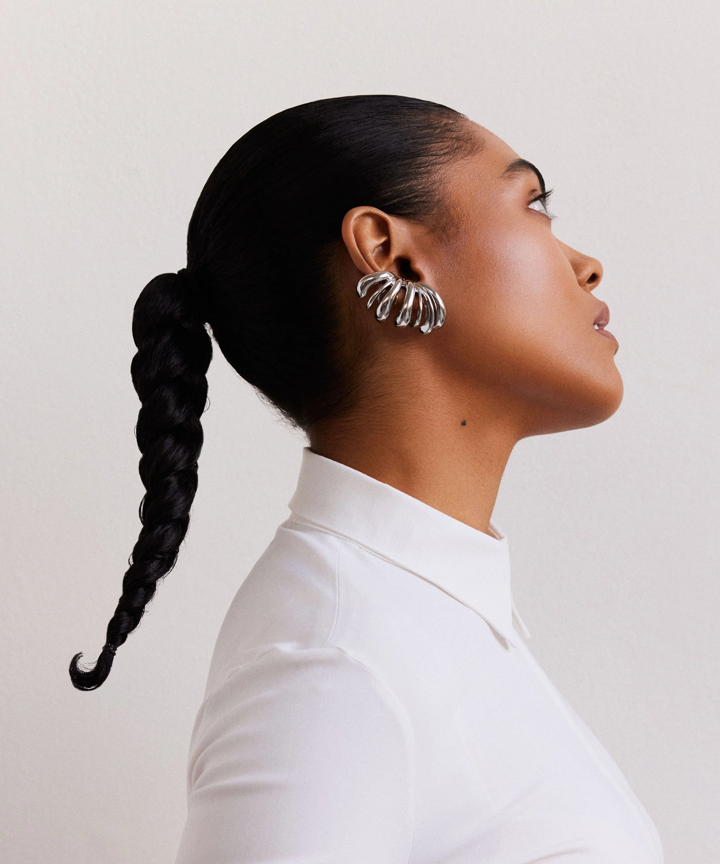 A woman with dark hair styled in a braid, wearing a white shirt and silver statement earring, posed in profile against a plain background.