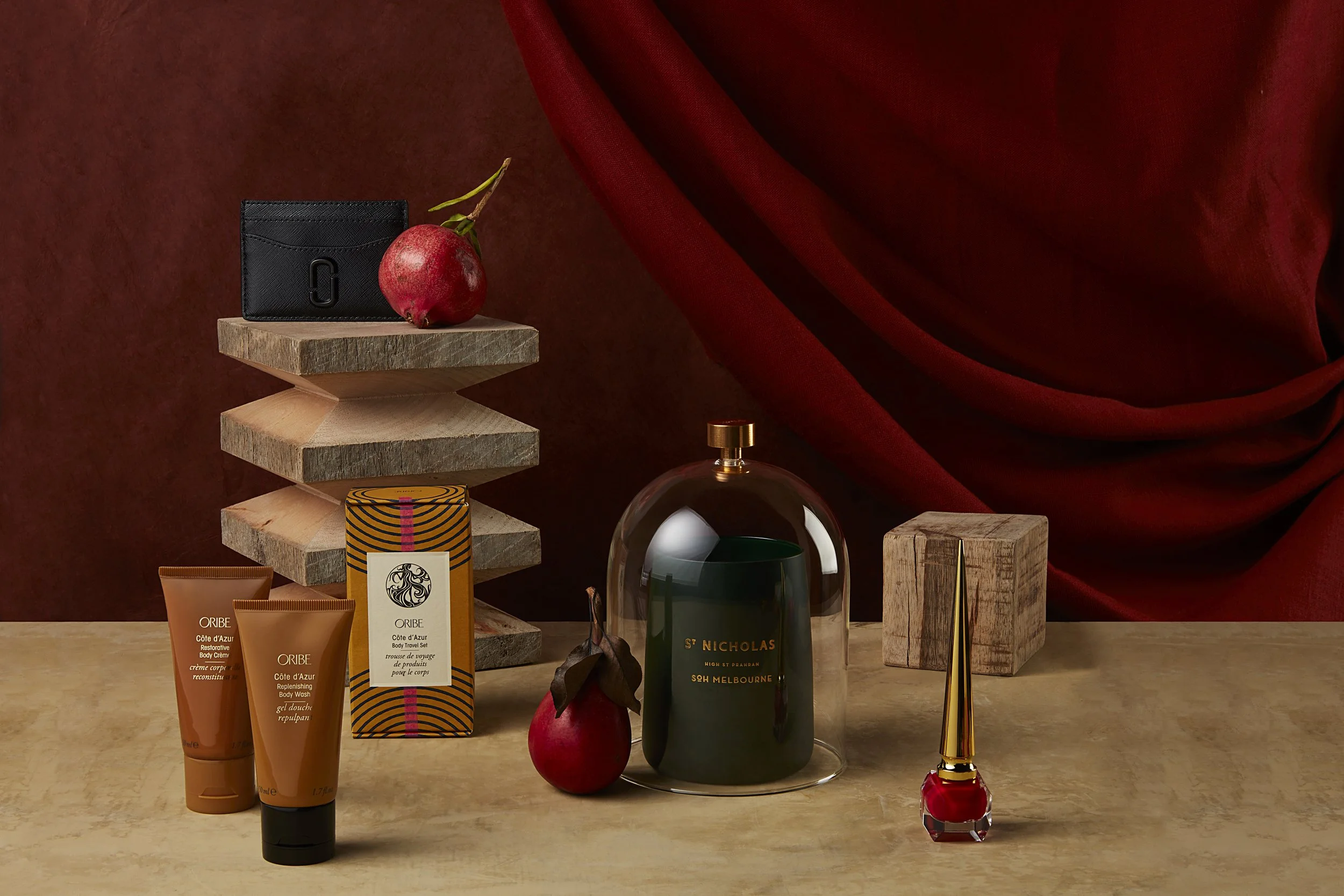 Arrangement of luxury items on a wooden surface, including body care products, a perfume bottle, a small wooden cube, a decorative glass dome, a red apple, and a red curtain in the background.