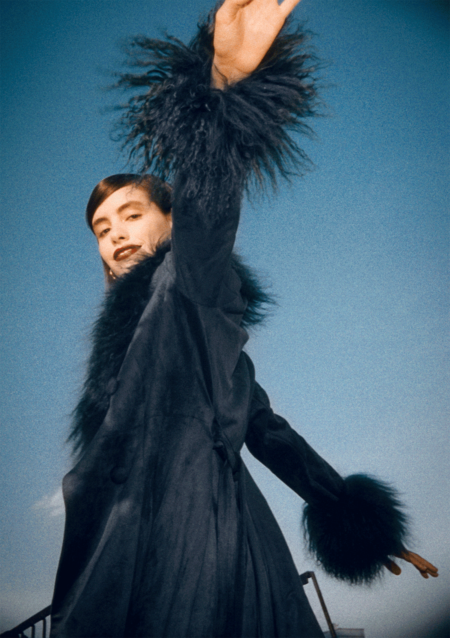 A woman with short dark hair is wearing a black coat with fluffy fur cuffs and trim, and is reaching out with her arm raised against a blue sky background.