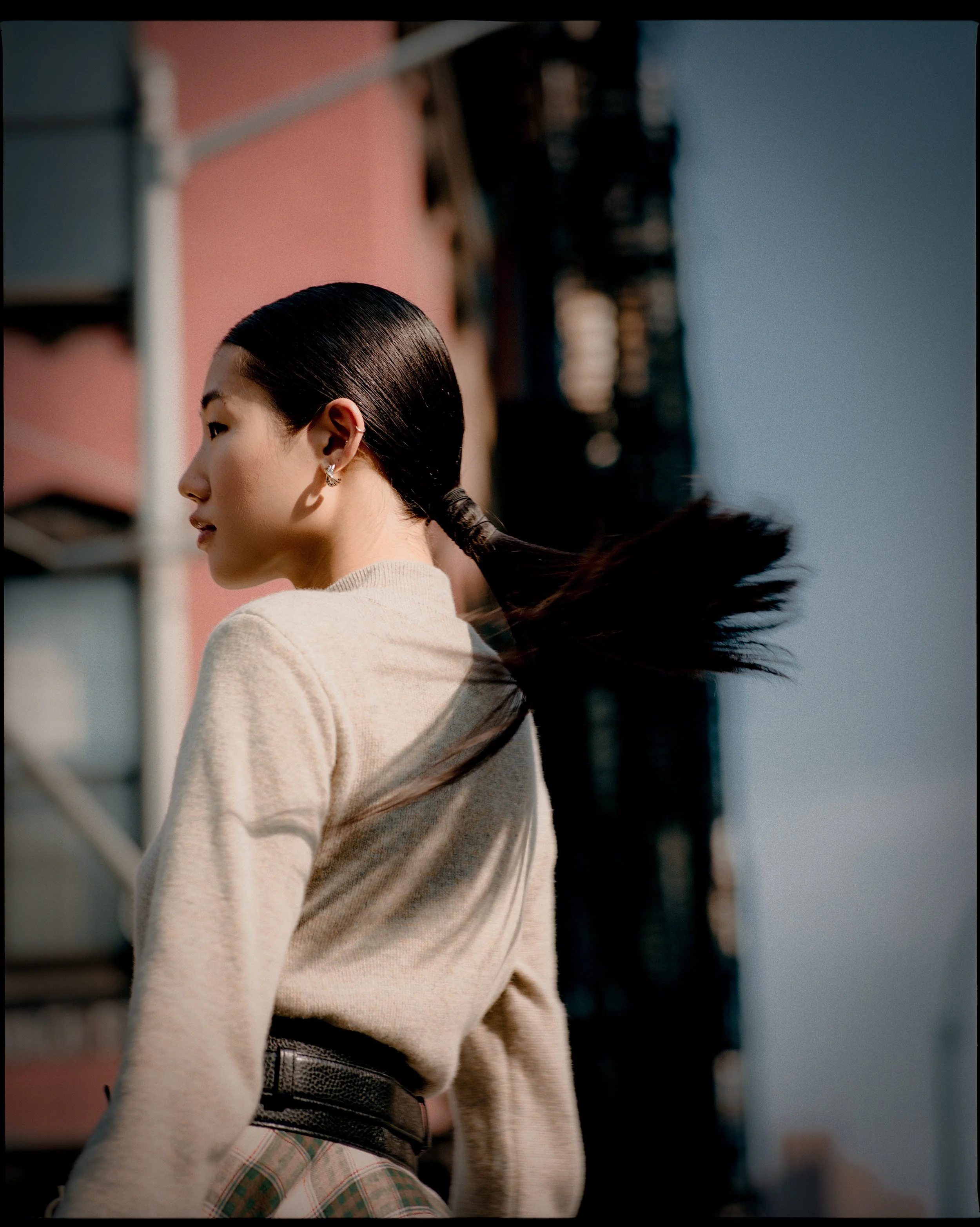 A young woman with dark hair tied in a low ponytail, wearing a beige sweater and plaid pants, stands outdoors with a cityscape background under a clear blue sky.