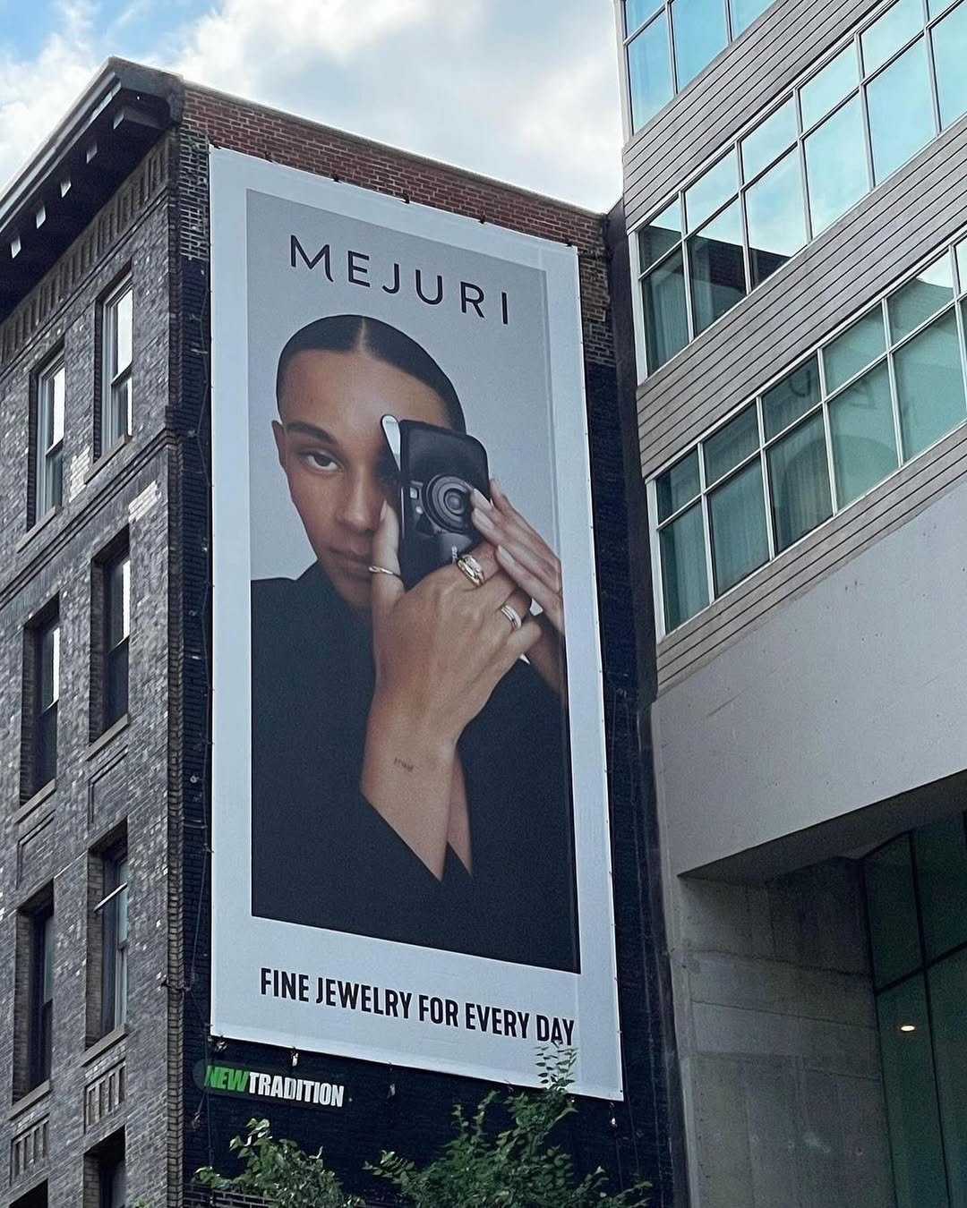 Large billboard featuring a woman holding a compact camera, with the text 'MEJURI' at the top and 'FINE JEWELRY FOR EVERY DAY' at the bottom, mounted on the side of a building in an urban area.