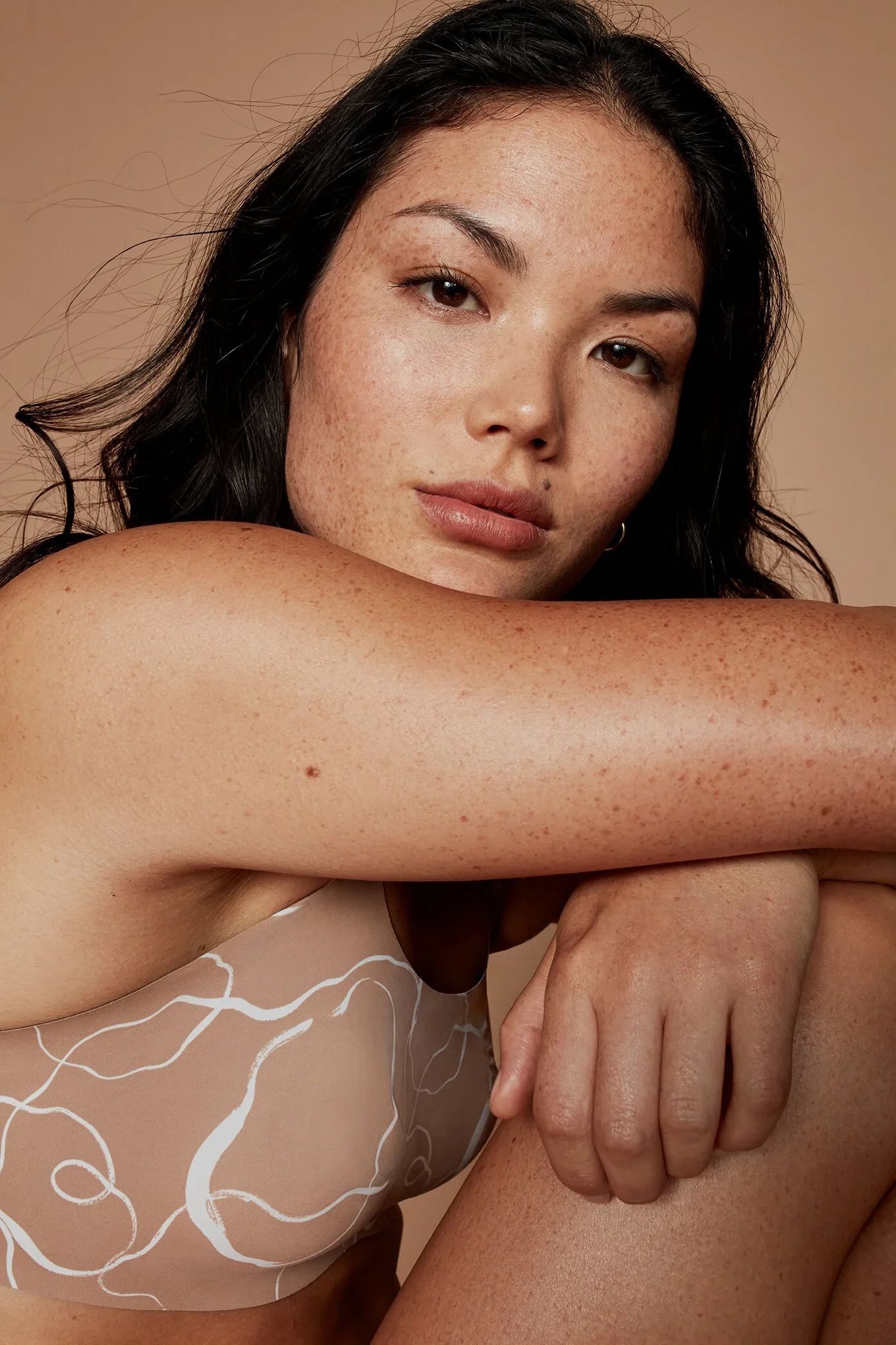 Close-up of a woman with dark hair and freckles, resting her arm over her knee, wearing a sheer top with white abstract line patterns against a neutral background.