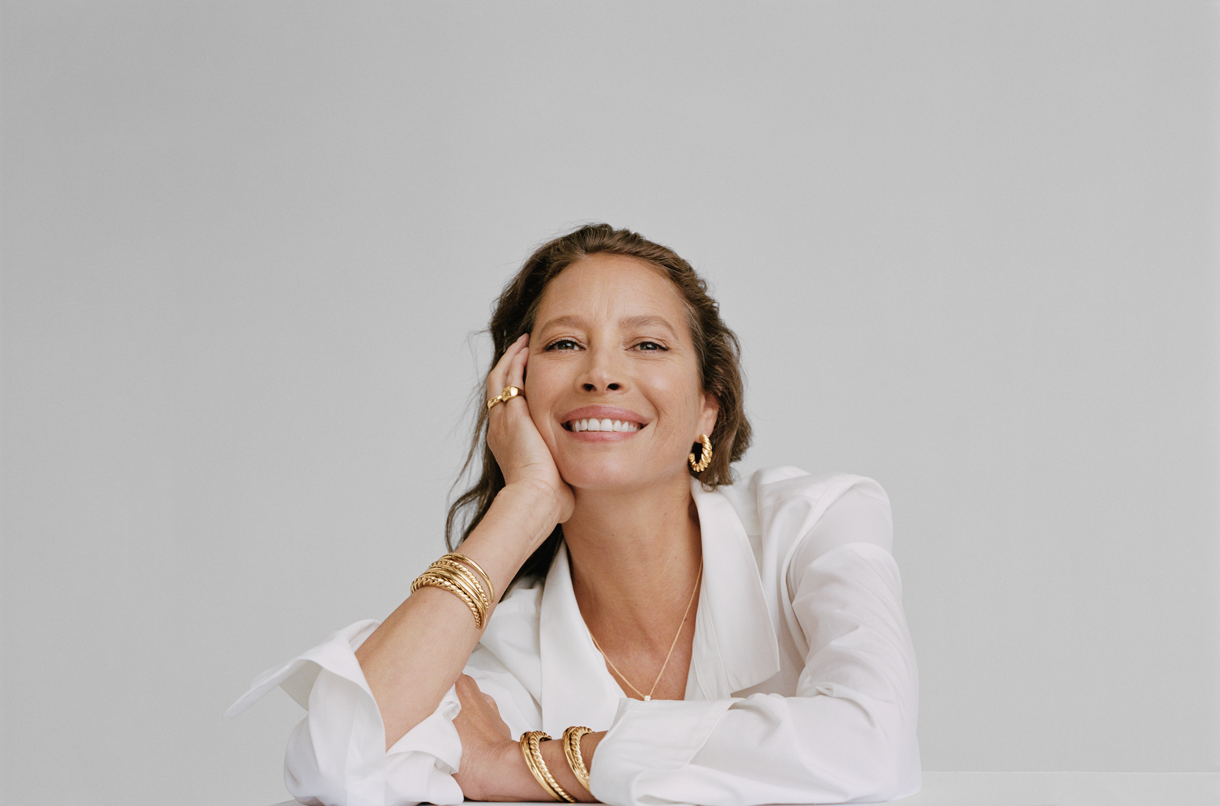 Portrait of a woman with short curly hair, wearing a white blazer and gold jewelry, smiling with her head resting on her hand against a plain light gray background.