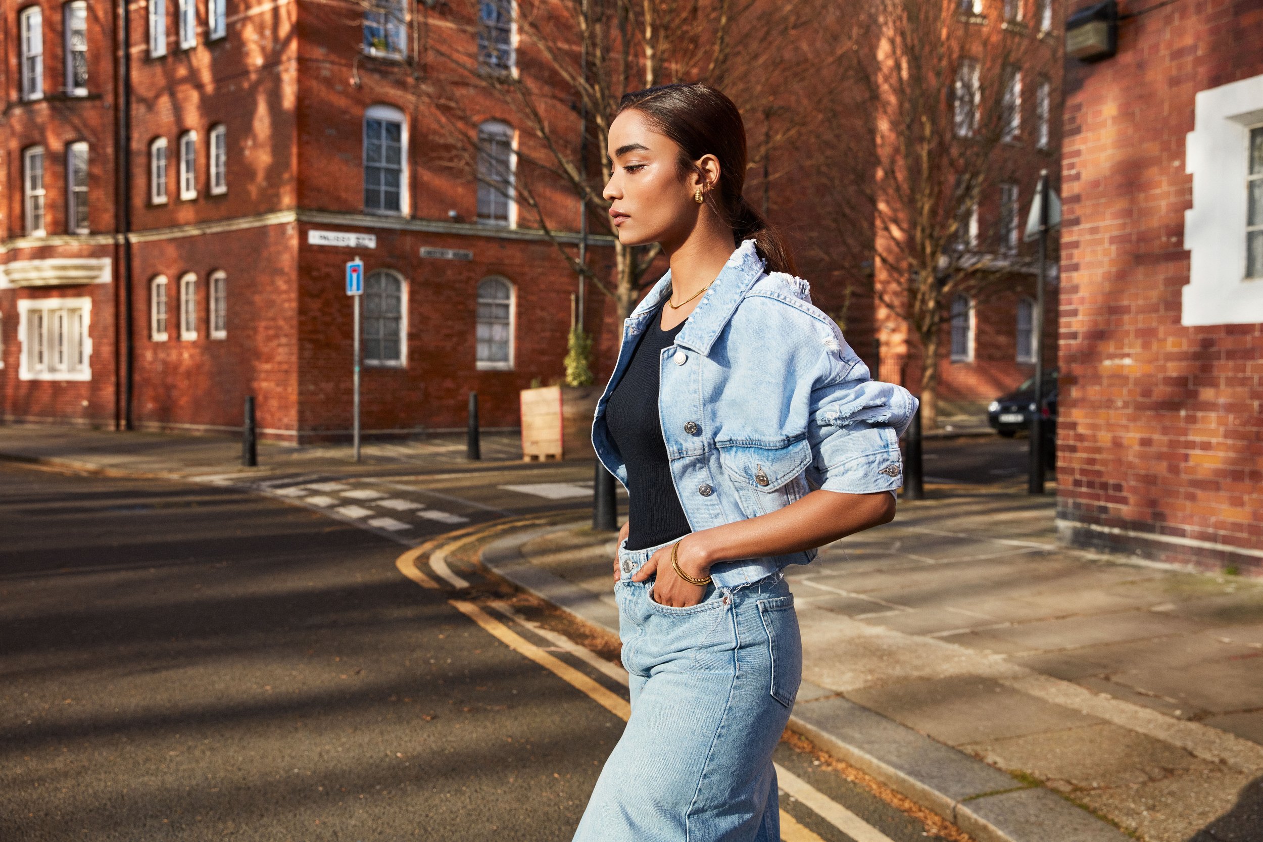 A young woman walking on a city street in autumn, wearing a light denim jacket, matching denim jeans, and gold accessories, with red brick buildings and leafless trees in the background.