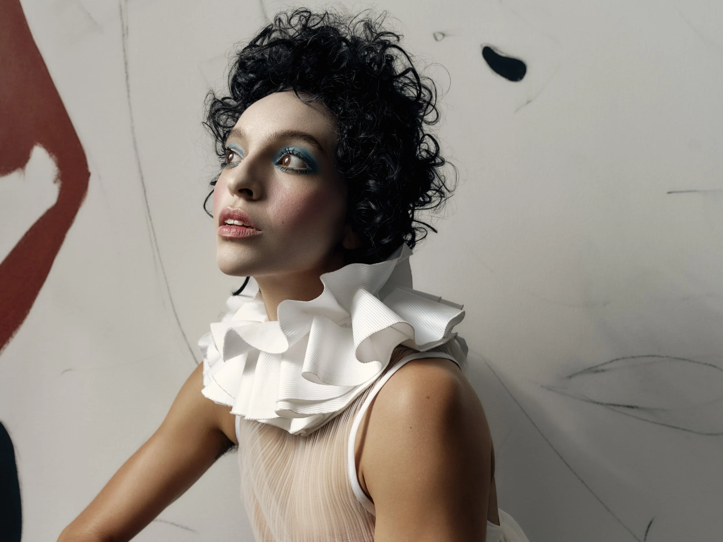 A woman with curly black hair and blue eye makeup wearing a white ruffled collar and a sleeveless top posing against a painted wall with abstract designs.