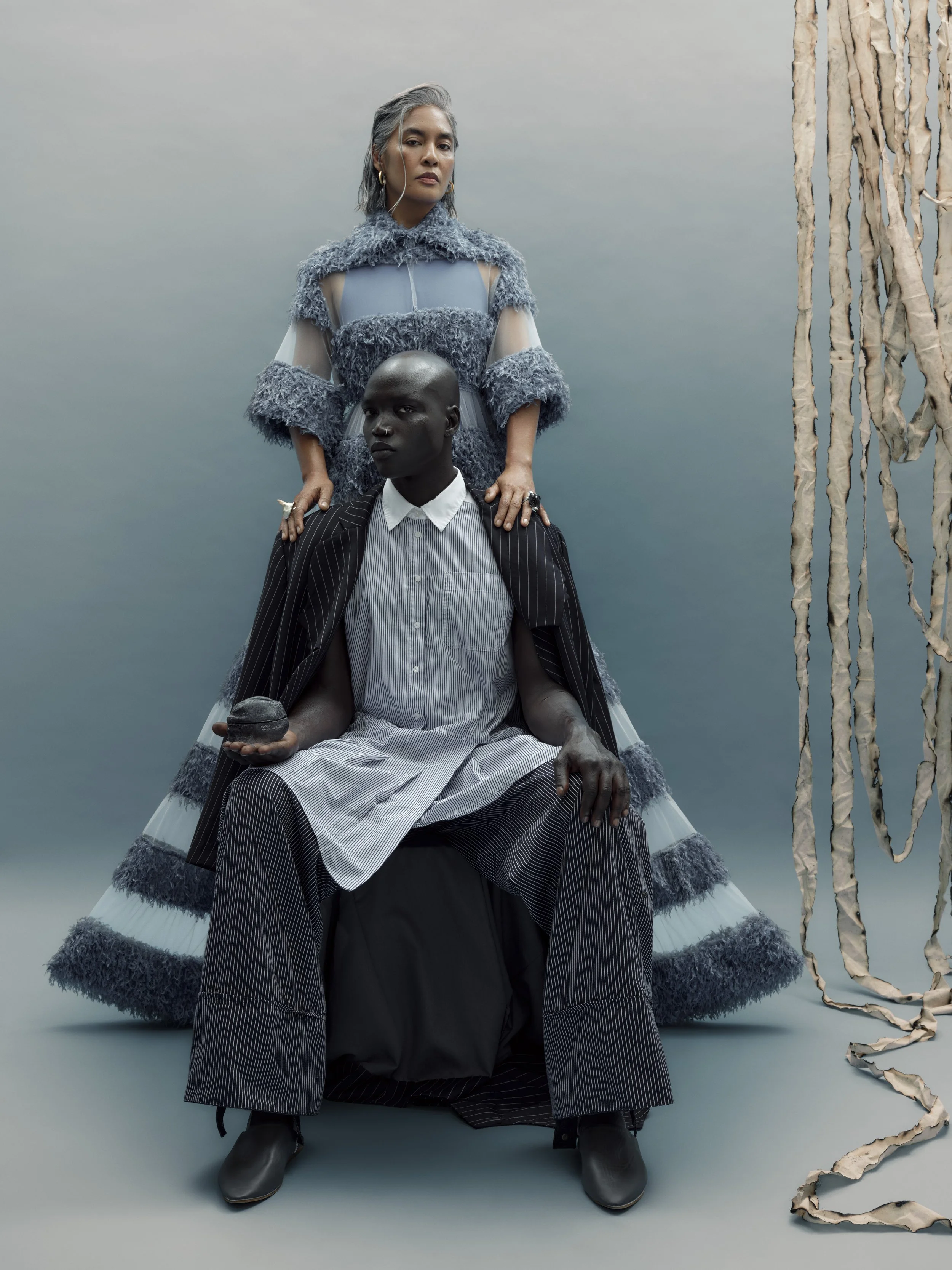 An artistic portrait of two fashion models against a somber, cloudy backdrop. The standing woman has silver hair, wearing a textured blue dress with sheer sleeves and fur accents. The seated man is dressed in a striped suit with a white shirt, holdin
