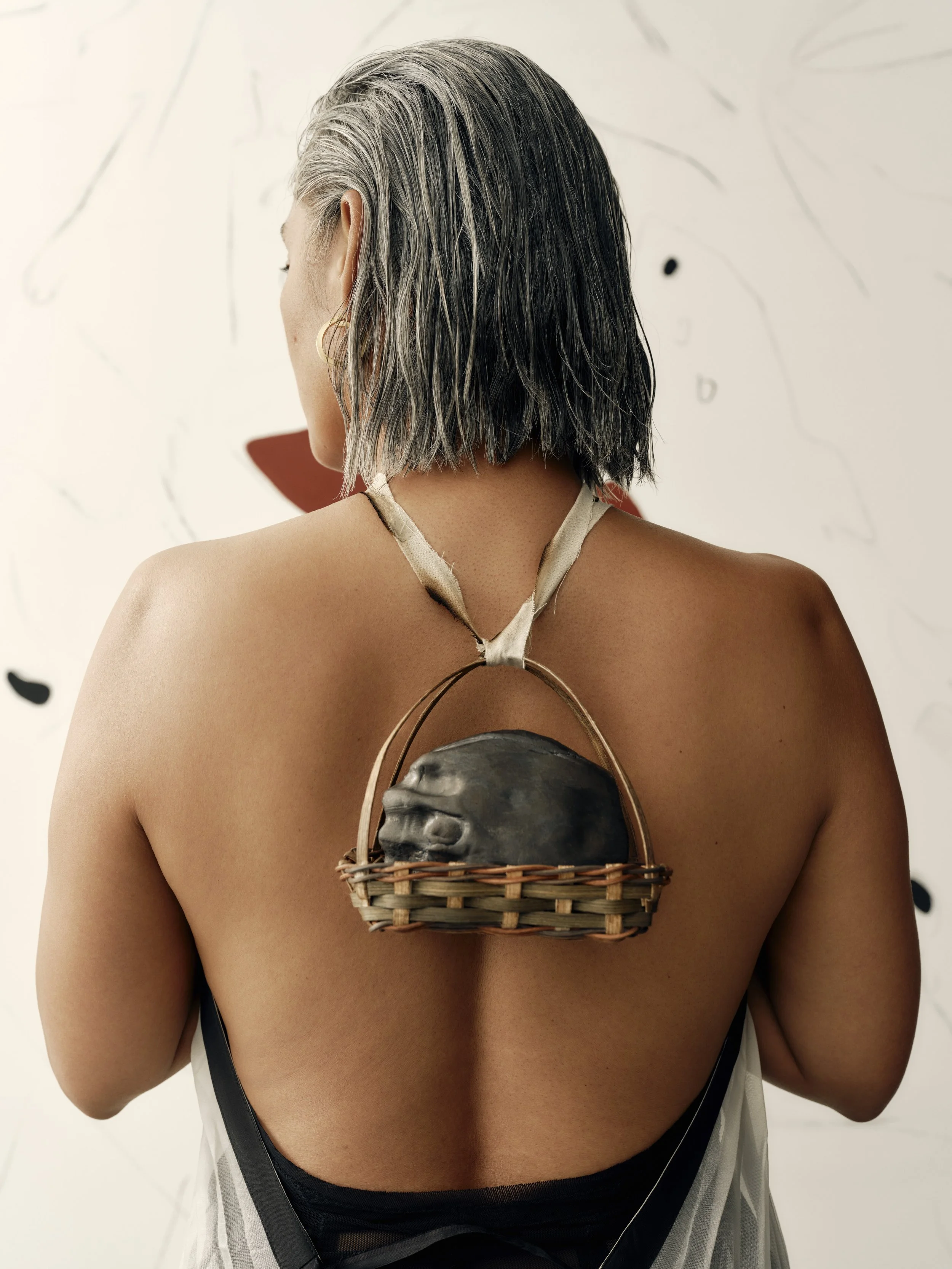 Back view of a woman with shoulder-length gray and black hair, wearing a halter top and a small basket on her back containing a rock or ceramic sculpture.