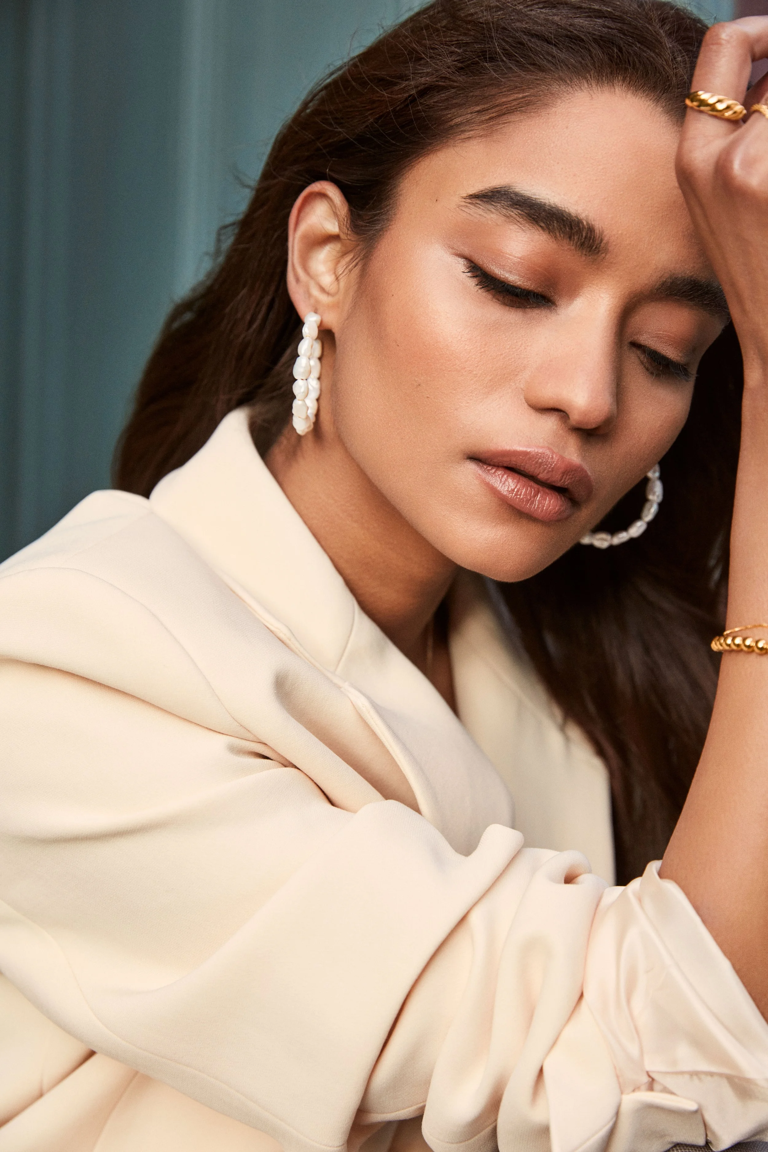 A woman with brown hair, wearing pearl earrings, a cream-colored blouse, and gold jewelry, resting her head on her arm with eyes closed.