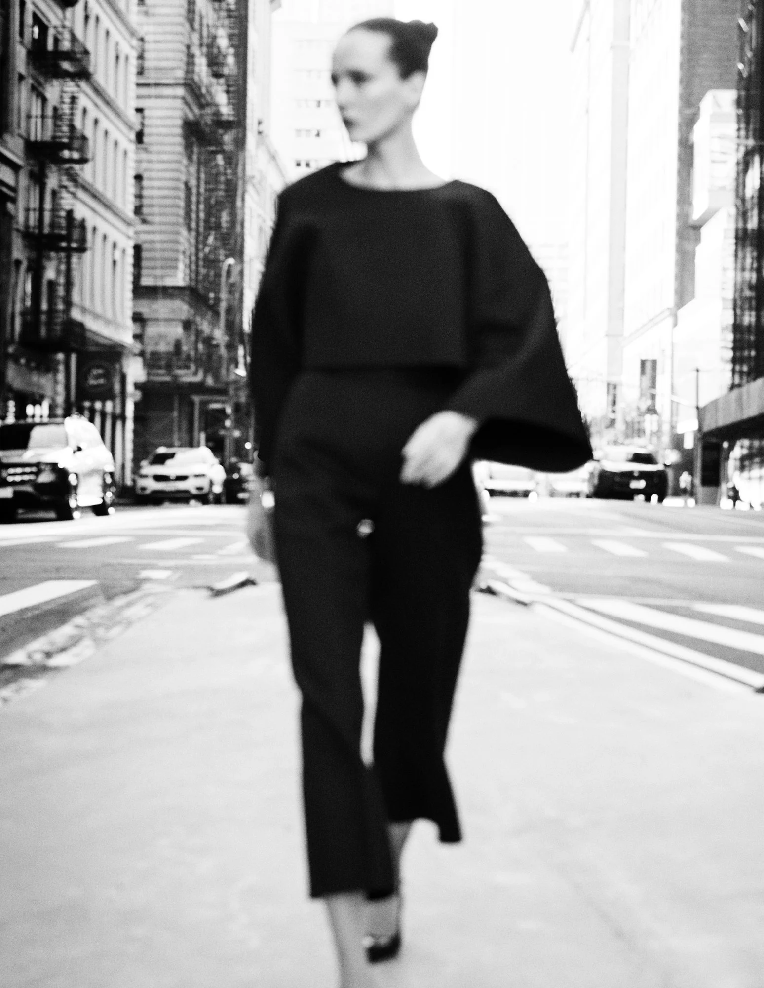 A woman walking on a city street in black and white, wearing a long-sleeved top and pants.