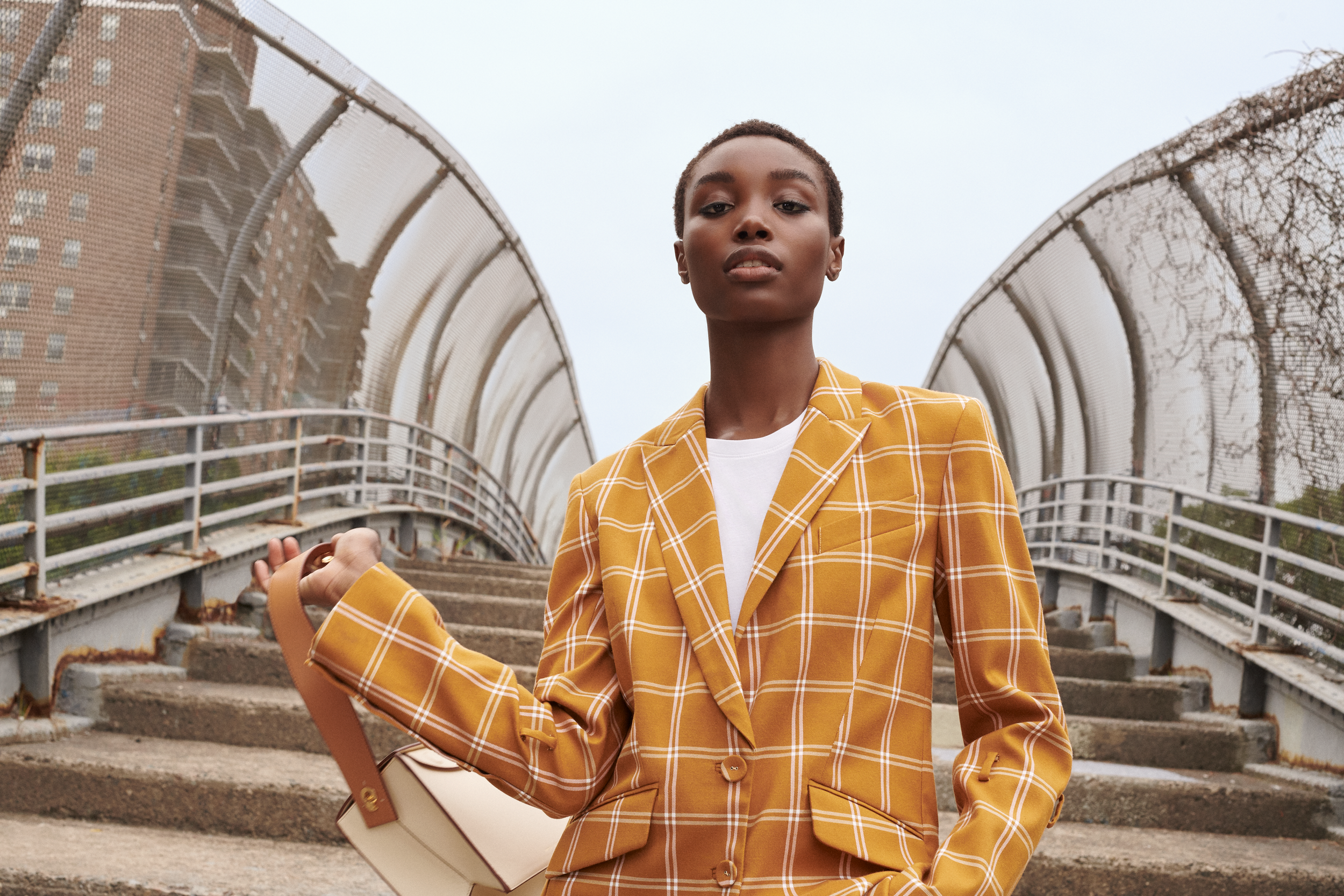 Person in a mustard plaid blazer standing on stairs outdoors, holding a beige handbag.