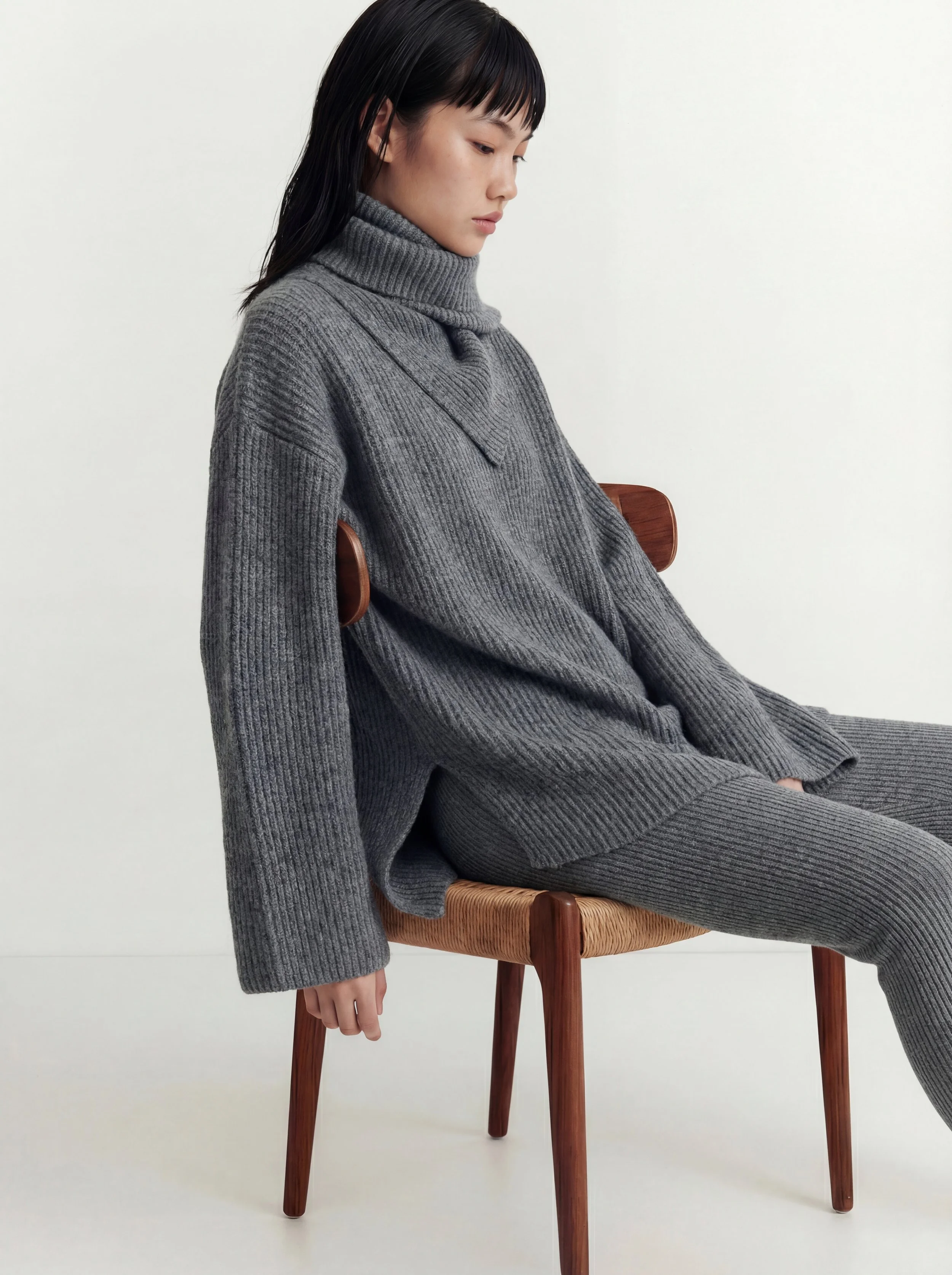 A woman in a gray knit turtleneck and matching pants sits on a wooden chair against a white background.