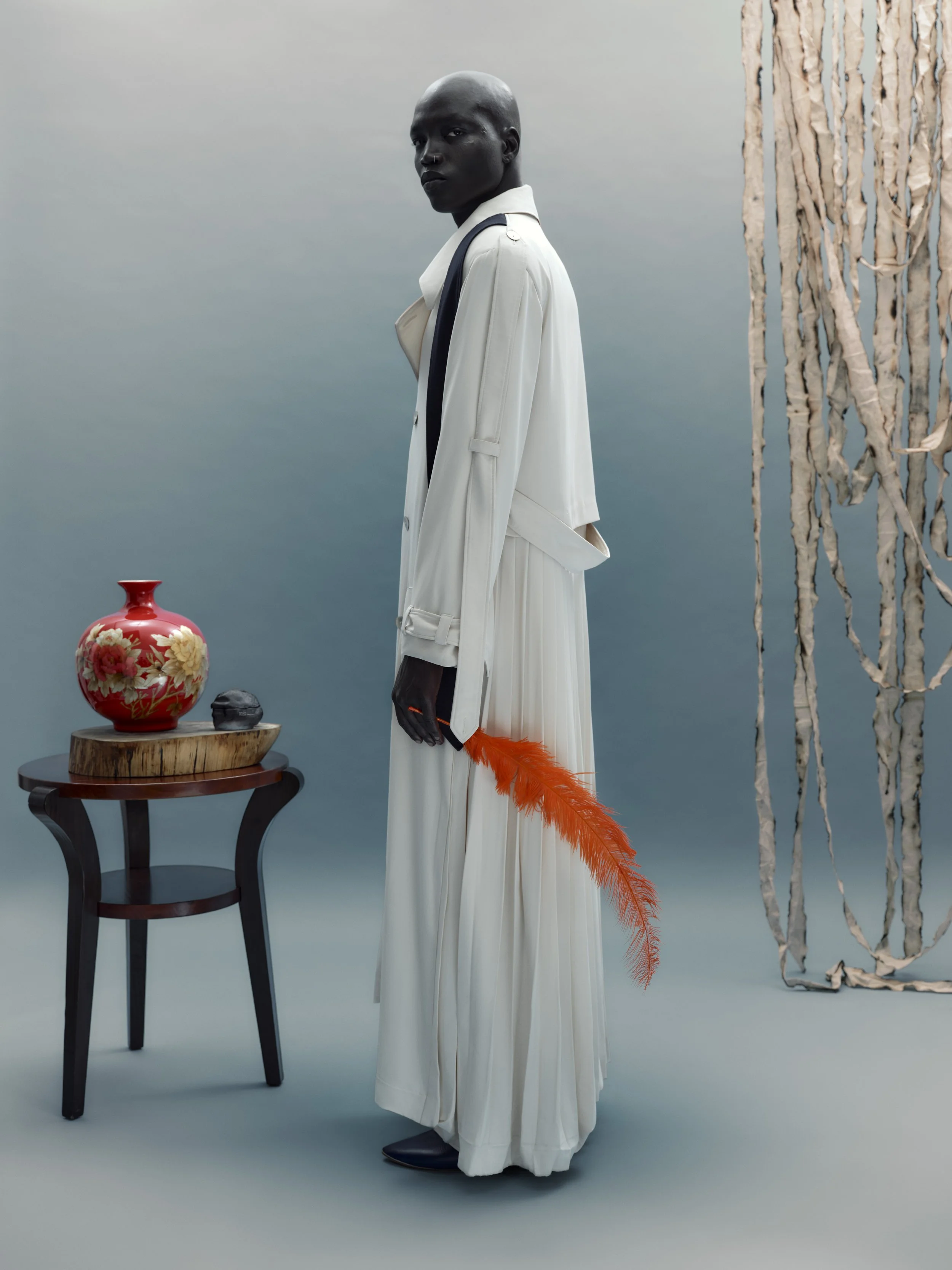 Fashion model with dark skin and shaved head dressed in a long white coat and pleated white dress, holding an orange feather in one hand, standing next to a wooden table with a red floral vase and sculpture inside, against a minimalist background wit
