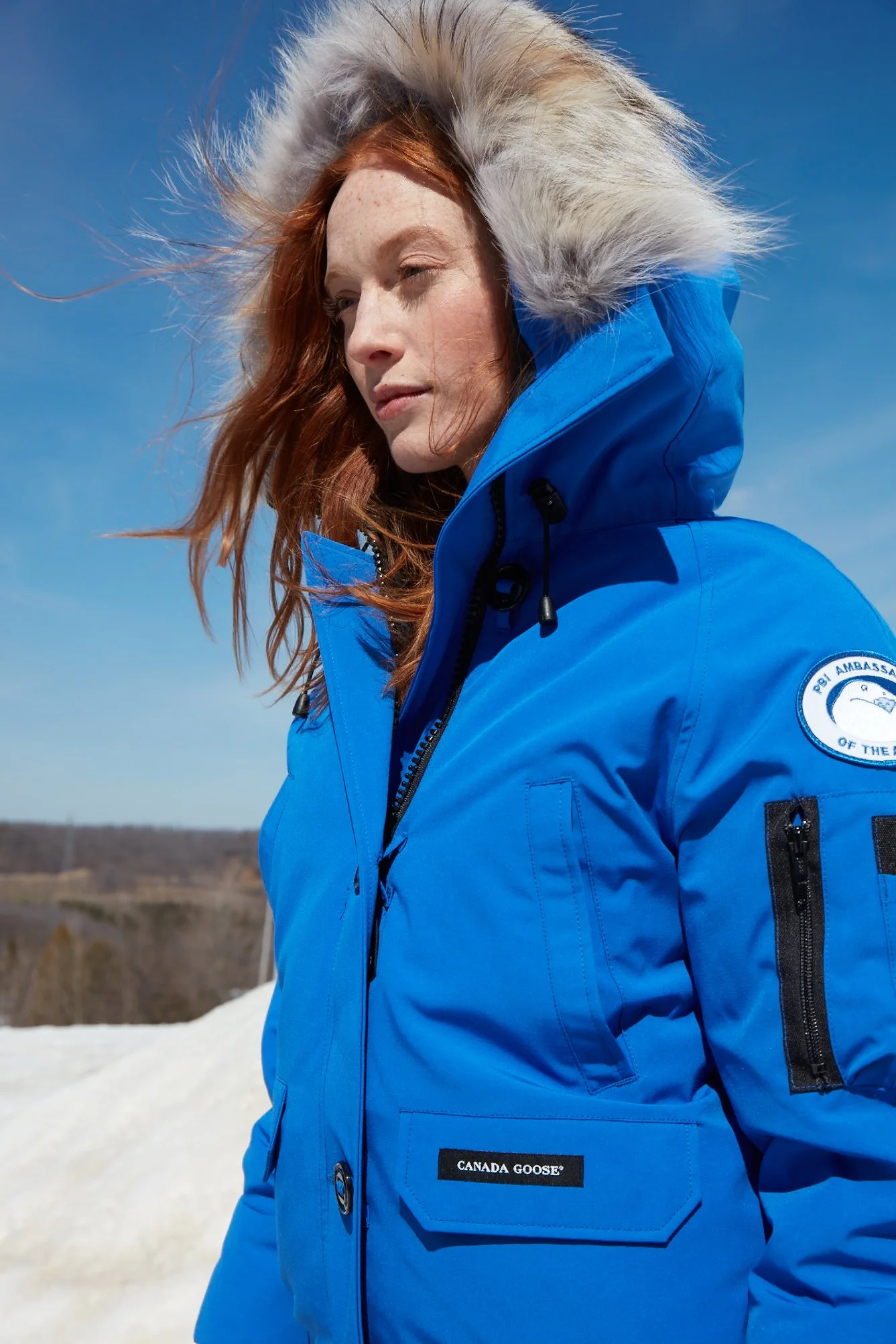 A woman with red hair wearing a blue Canada Goose winter jacket with a fur-lined hood outdoors on a clear day.