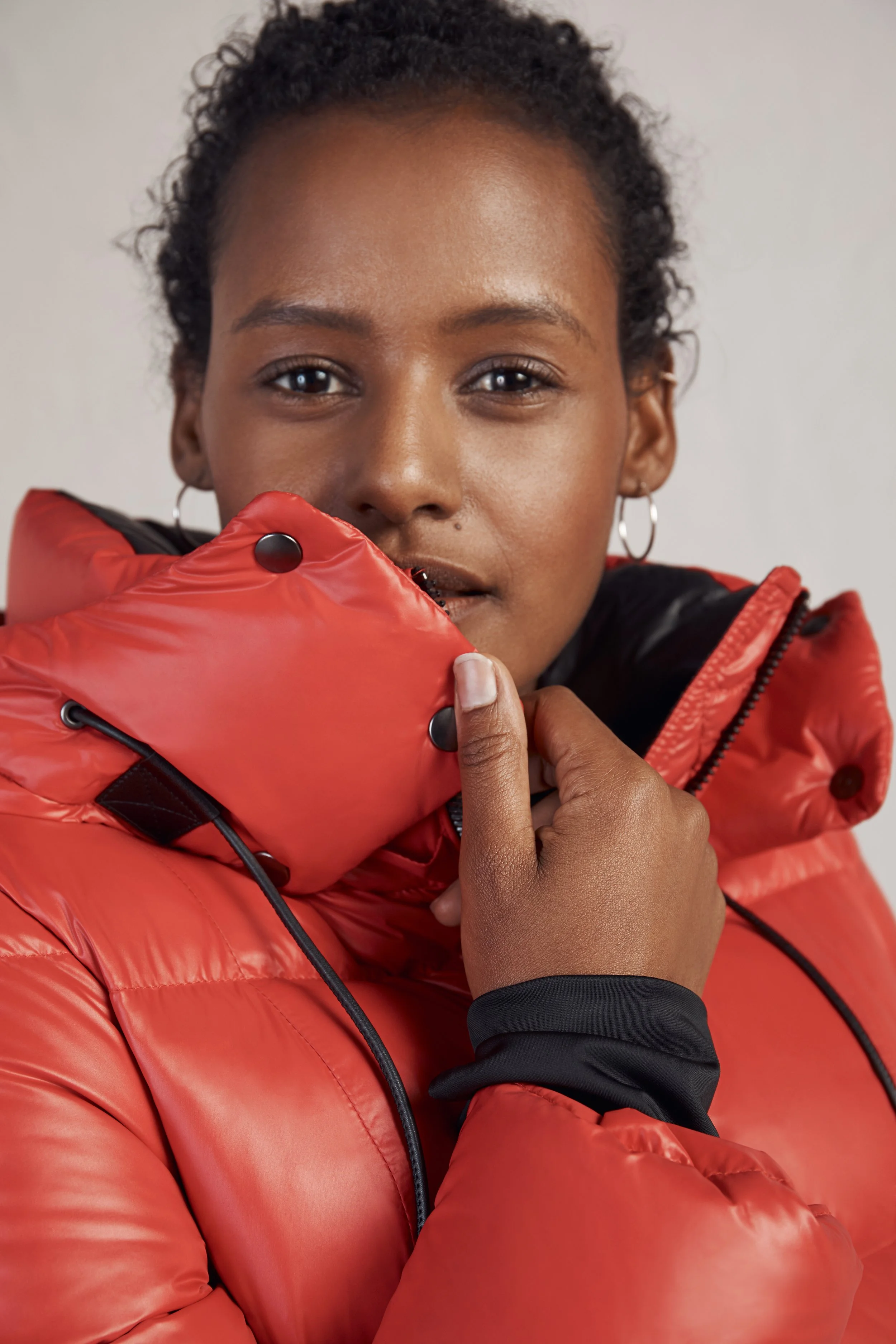 A woman wearing a red puffer jacket with a black inner lining holds the collar of her jacket up to her face. She has short curly hair and is wearing small hoop earrings, with a neutral expression and looking directly at the camera.