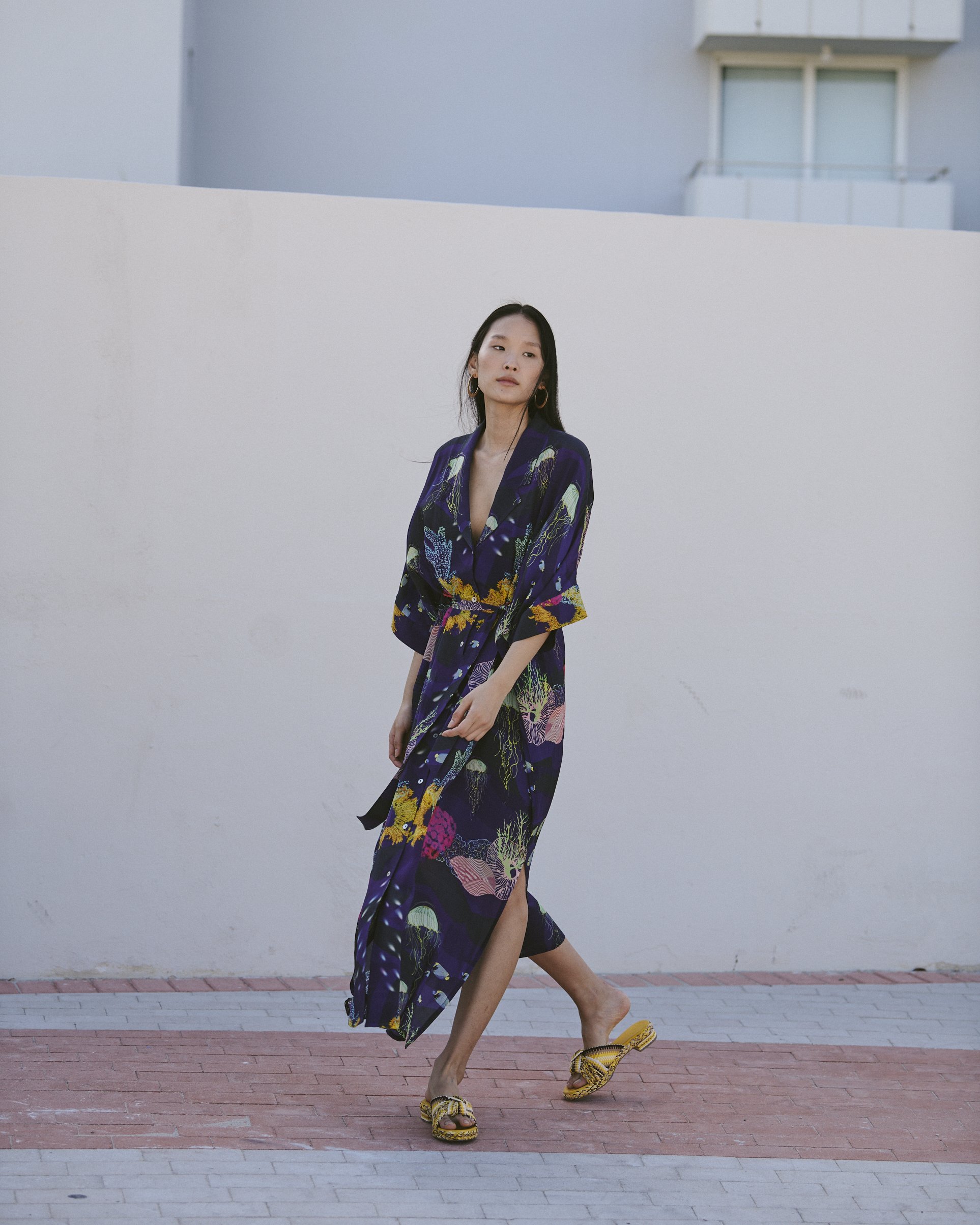 A woman walking on a brick sidewalk, wearing a long, dark dress with a floral pattern and yellow sandals, against a plain white wall.