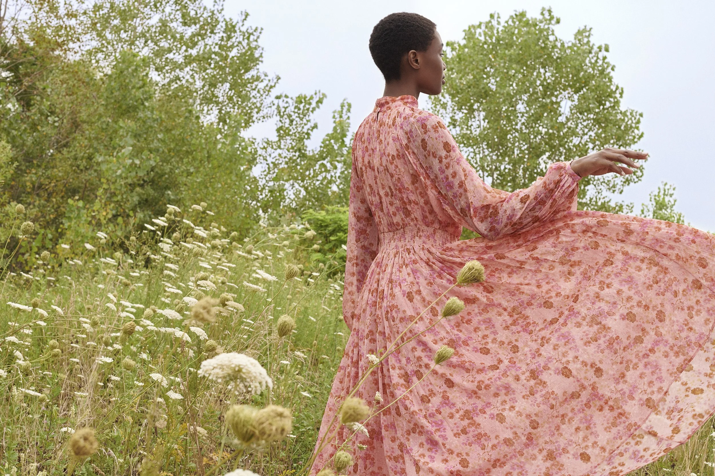 A woman in a pink floral dress sitting in a grassy field with trees in the background.