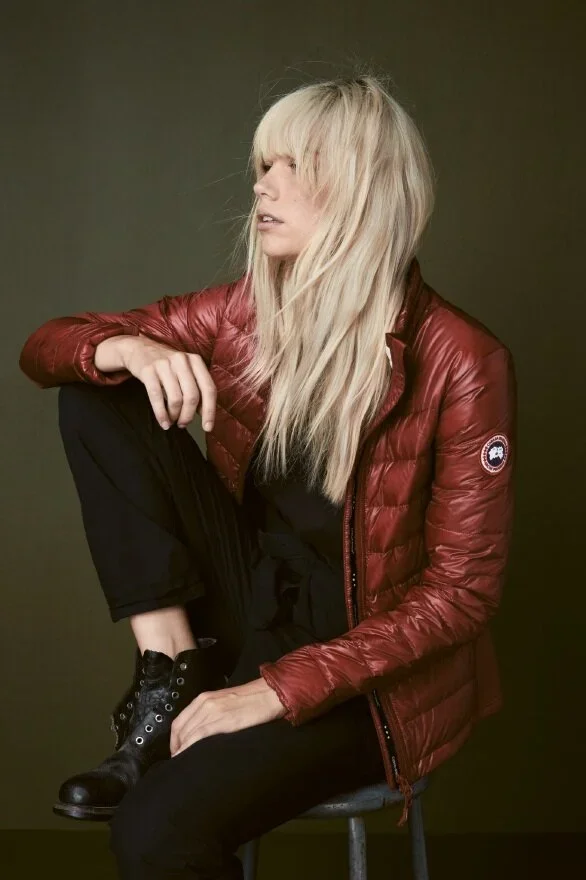 A woman with long, blonde hair wearing a red puffy jacket, black pants, and black boots, sitting on a stool against a dark background.