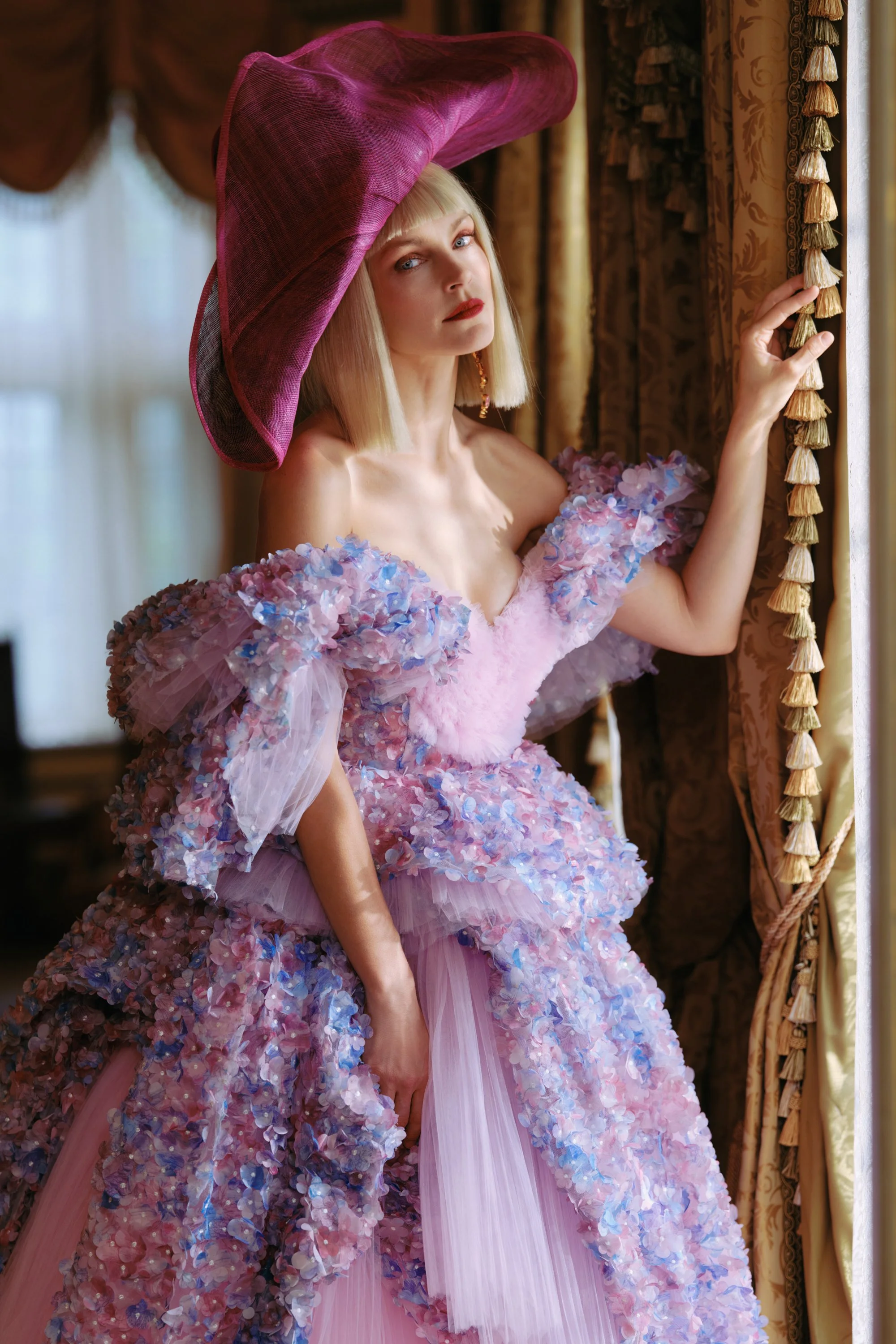A woman wearing a purple, floral, ruffled ball gown and a large purple hat, standing by a window with ornate curtains.
