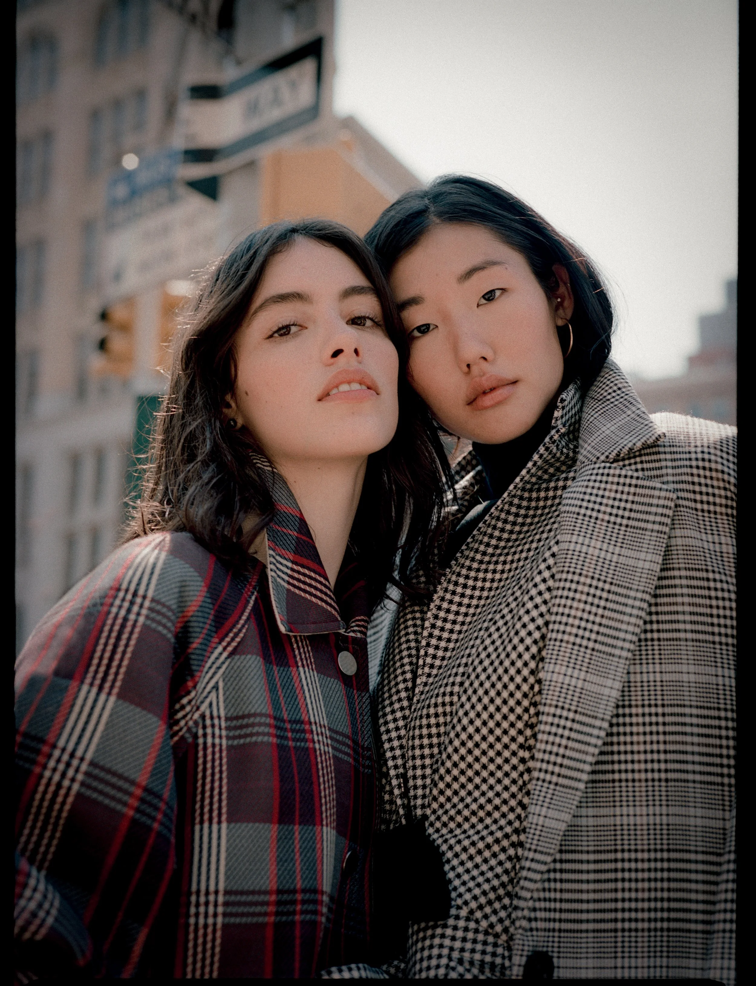 Two women standing close together outdoors, wearing plaid coats, with city buildings and street signs in the background.